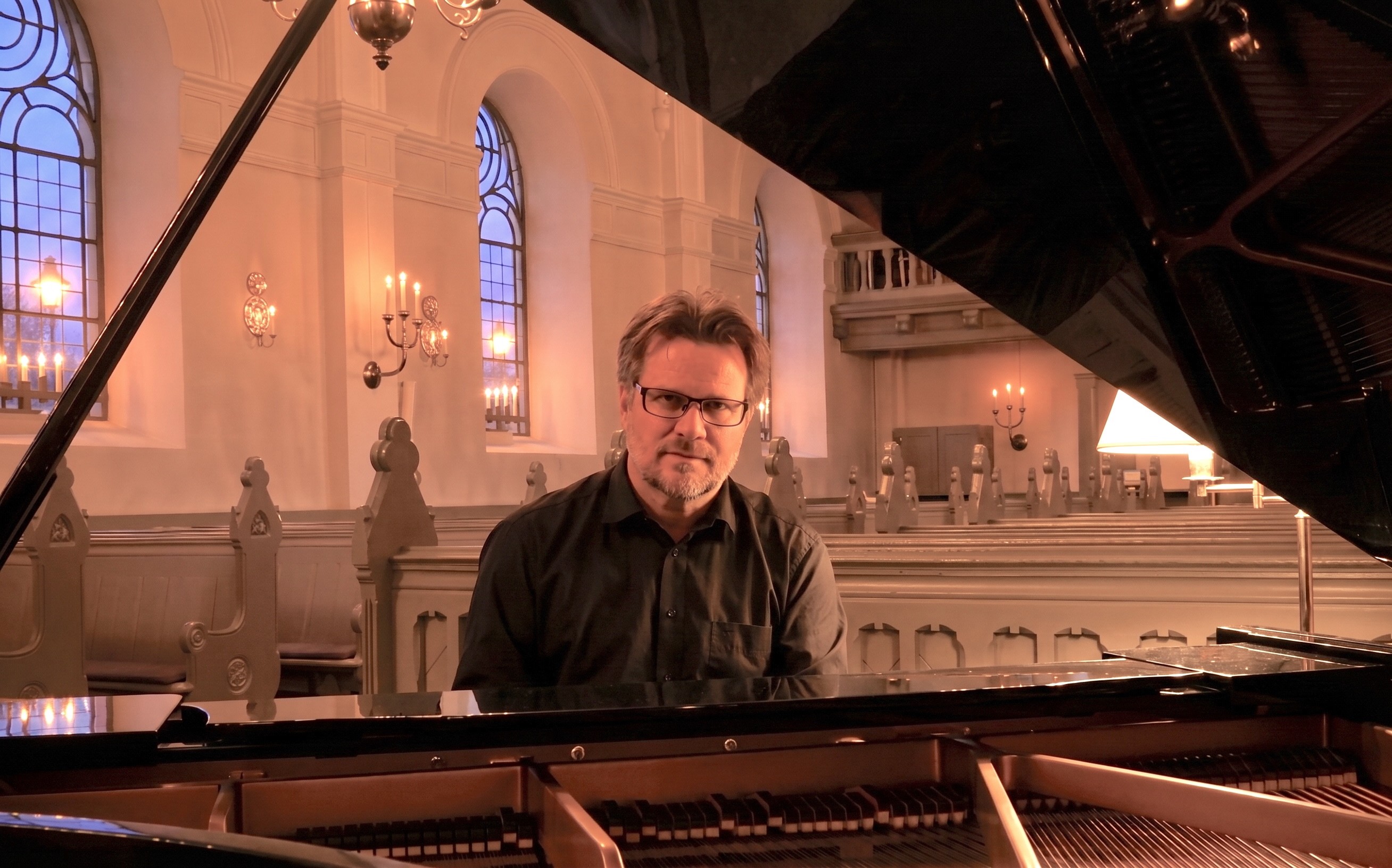 En man i mörk skjorta, glasögon och kortklippt brunt hår sitter vid en öppen flygel i en kyrka. Grågröna kyrkbänkar, vitputsade väggar och stora kyrkfönster i bakgrunden.