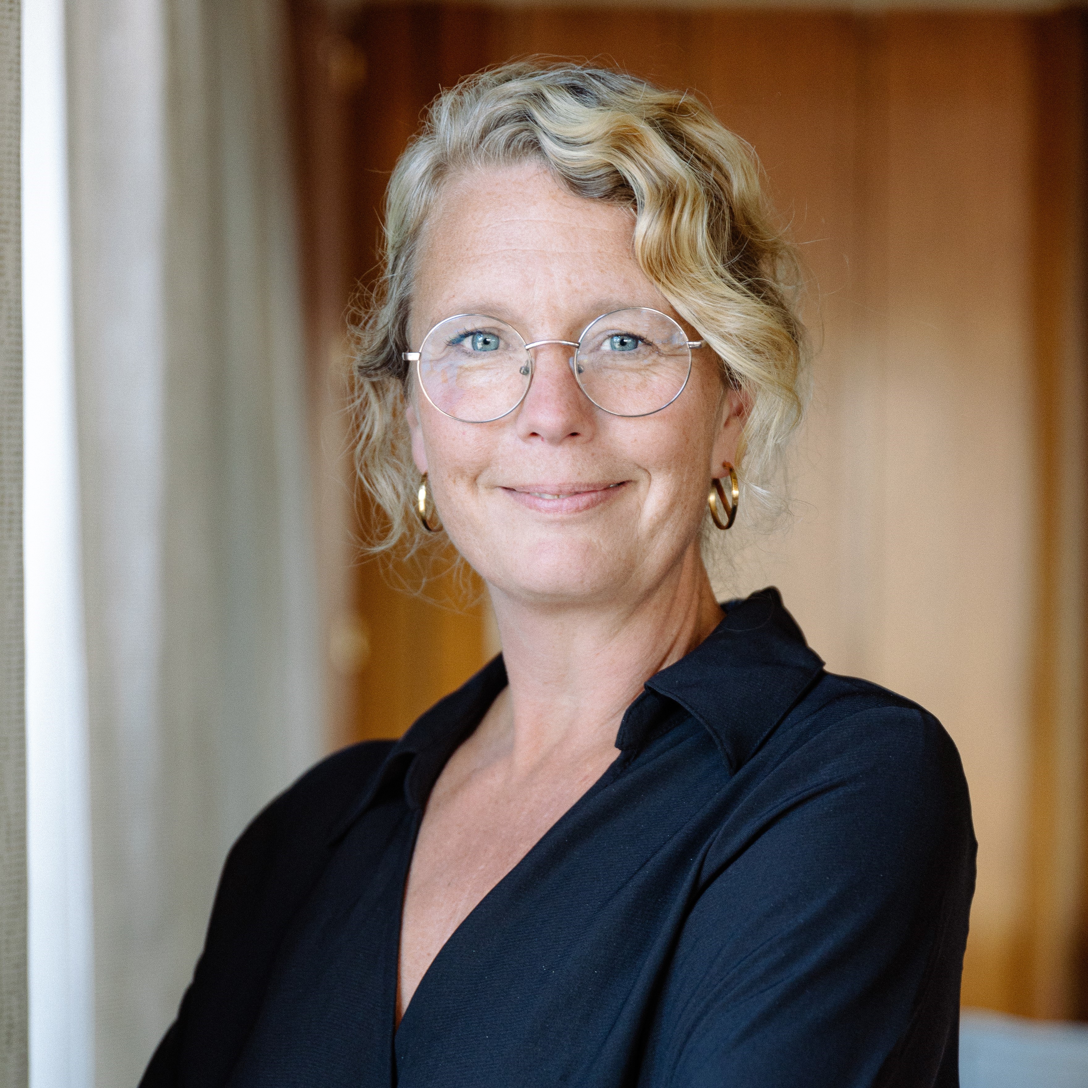 Leende kvinna med uppsatt, ljust, lockigt hår, guldbågade glasögon och guldringar i öronen. Hon bär en blå blus som är öppen i halsen.