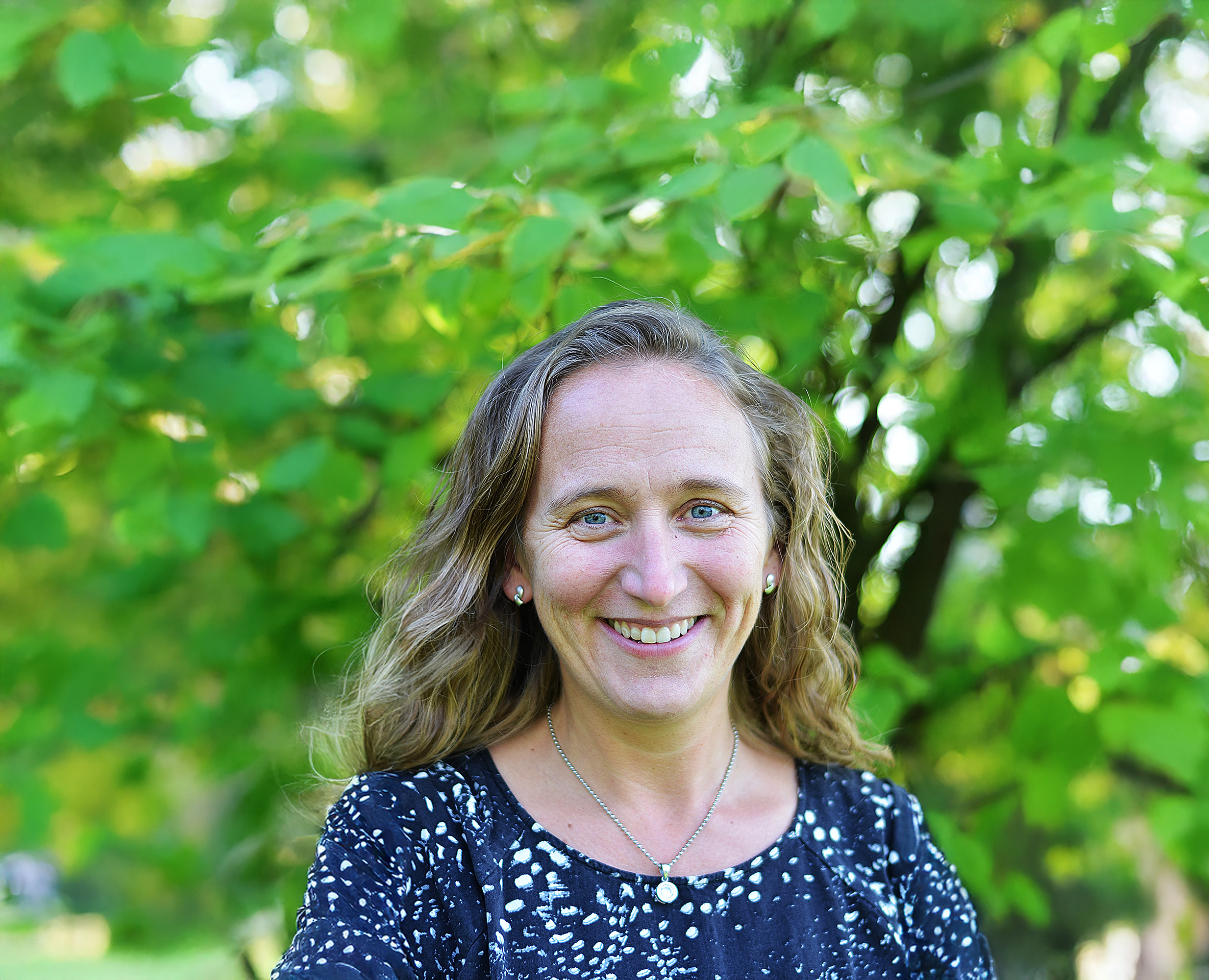 Leende kvinna med brunt, långt, lockigt hår och blå blus. Gröna blad i bakgrunden.
