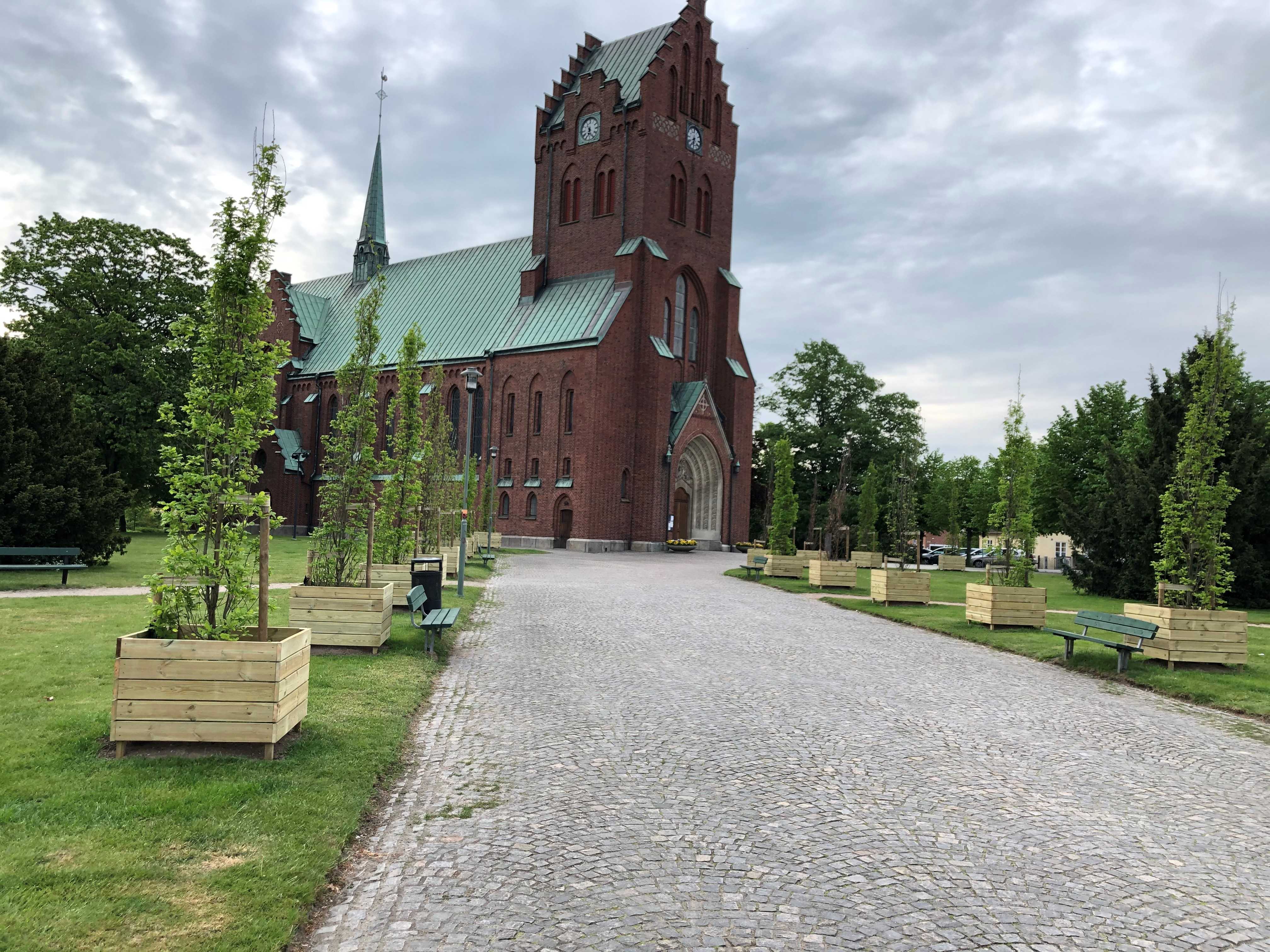 Bred gång kantad av pelarekar framför Hässleholms ståtliga kyrka i rött tegel.