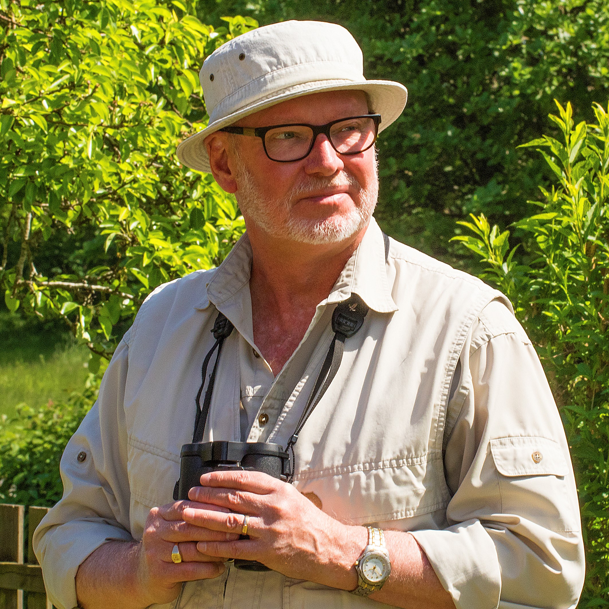 Man med ljust skägg och svartbågade glasögon i ljus solhatt och ljus skjorta i solen framför träd med starkt gröna blad.