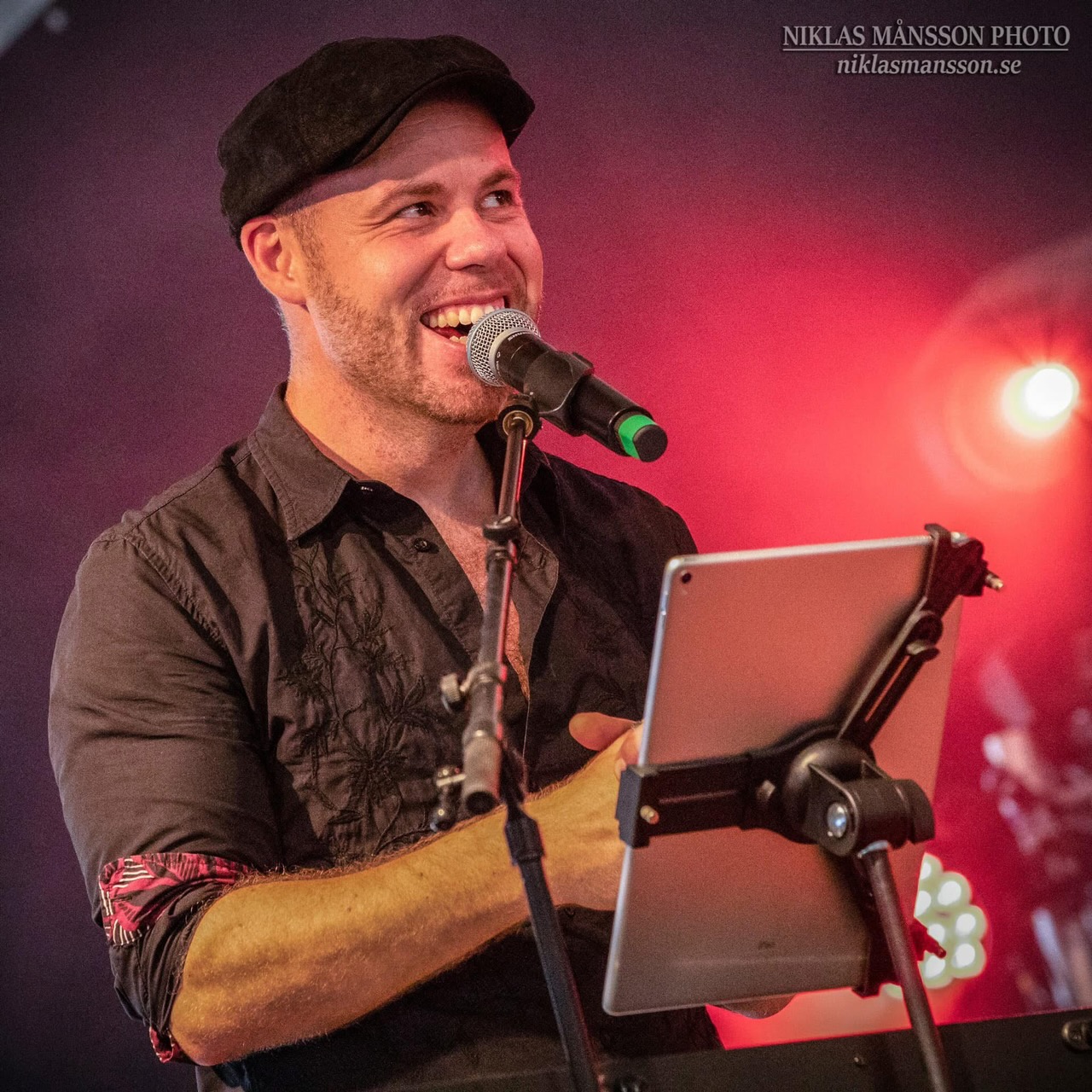 En man med keps och lite skägg sjunger i en mikrofon. Rödaktig bakgrund.