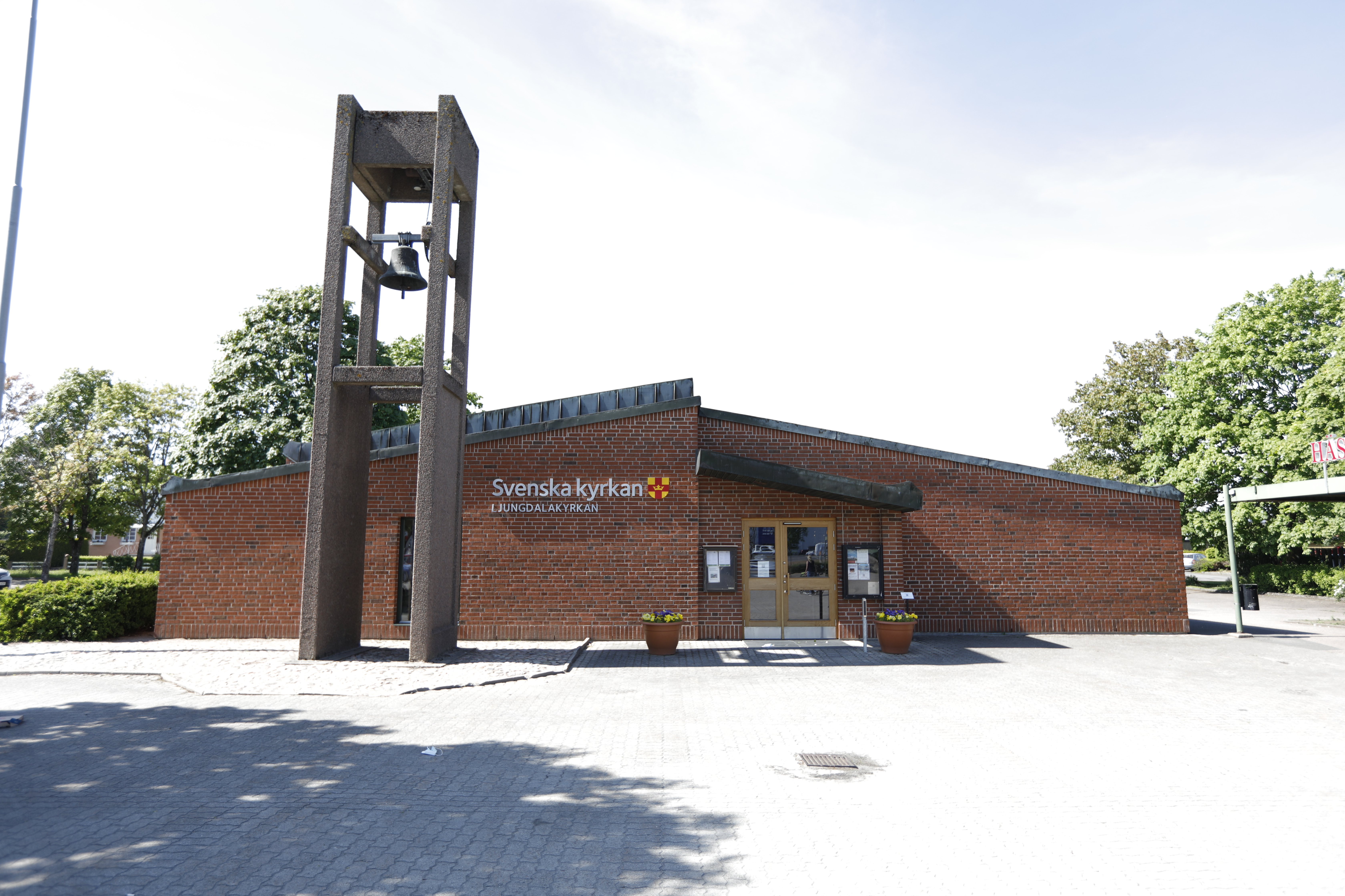 Brunröd tegelkyrka med klockstapel framför. Gröna träd vid sidorna.