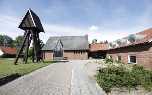 Krattad bred grusgång leder fram till brun tegelkyrka. Klockstapel av trä till vänster i bild. Grön gräsmatta och gröna buskar.