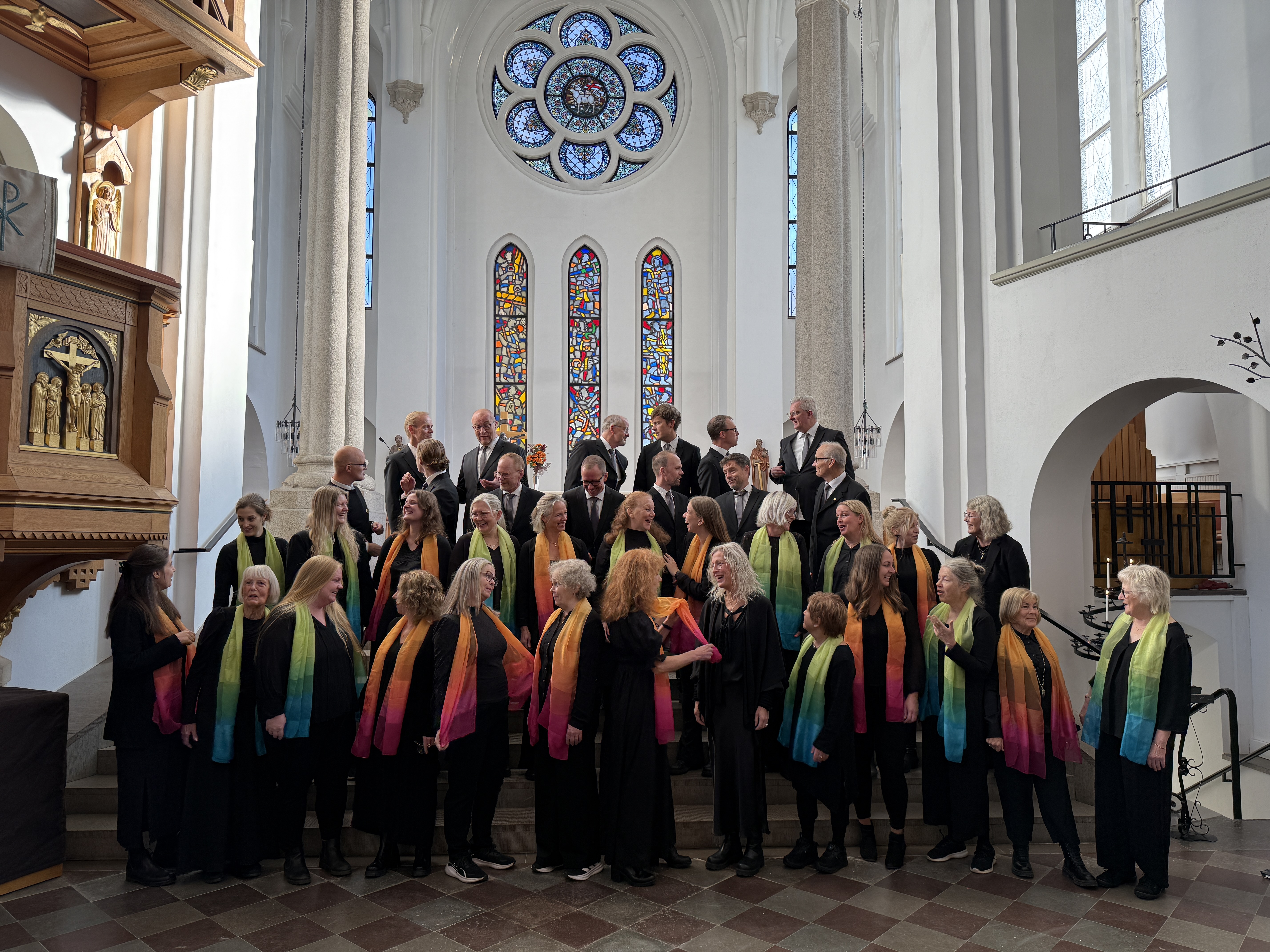 Svartklädda  män och kvinnor med färgglada sjalar står i kyrkans kor och samtalar med varandra i en paus i körövningen