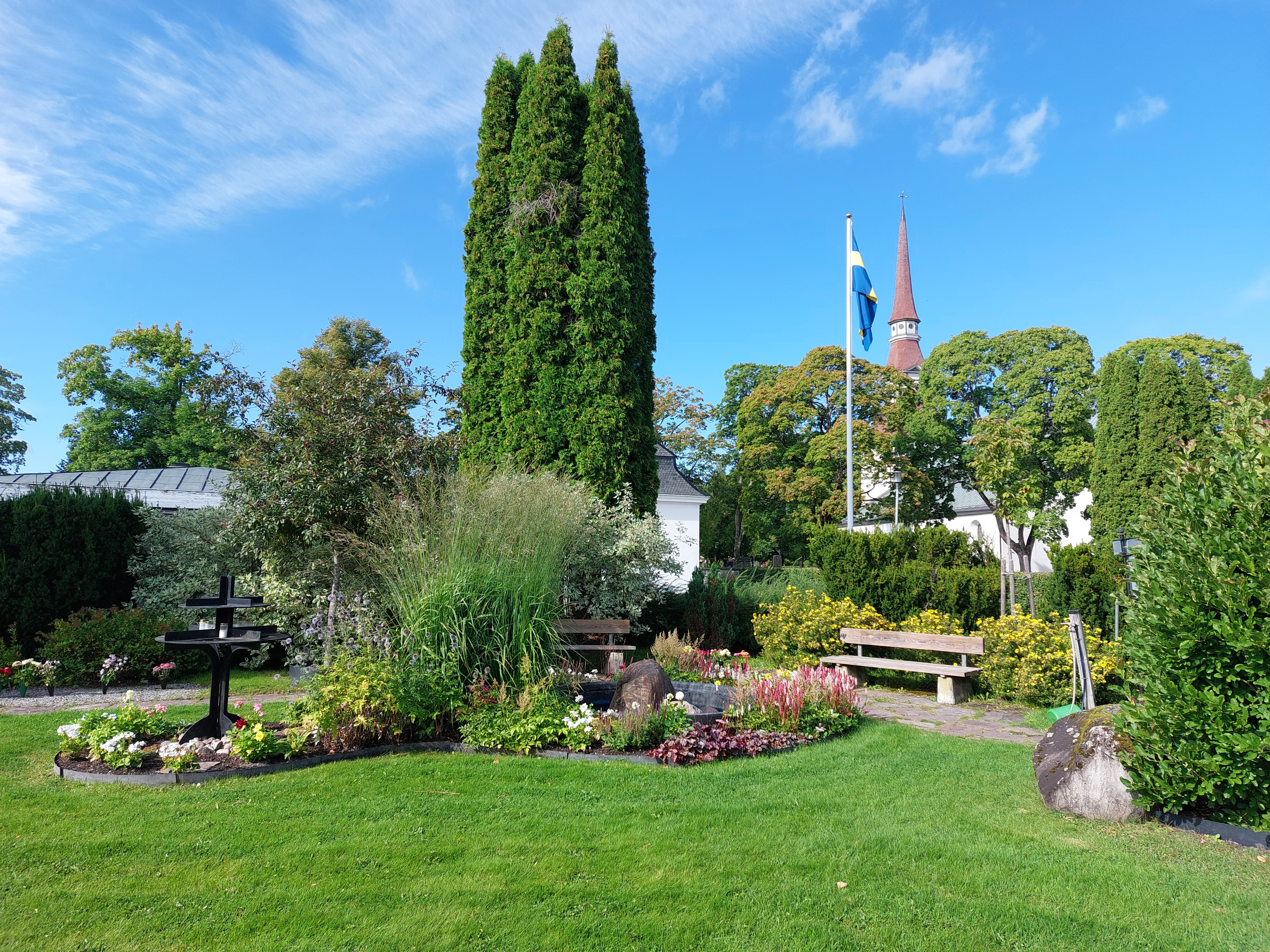 Minneslunden på kyrkogården i Norrbärke, en sommarbild med blommor och gröna träd. Norrbärke kyrka skymtar i bakgrunden.