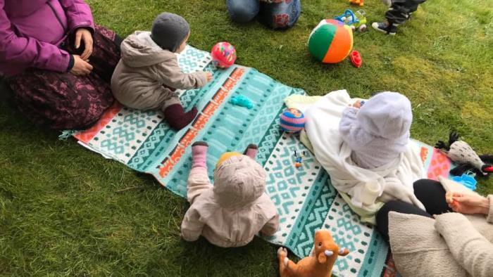 Barn och föräldrar sitter på en färgglad matta utomhus och leker. 