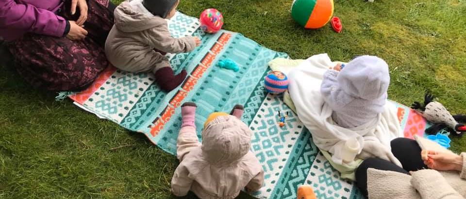 Barn och föräldrar sitter på en färgglad matta utomhus och leker. 