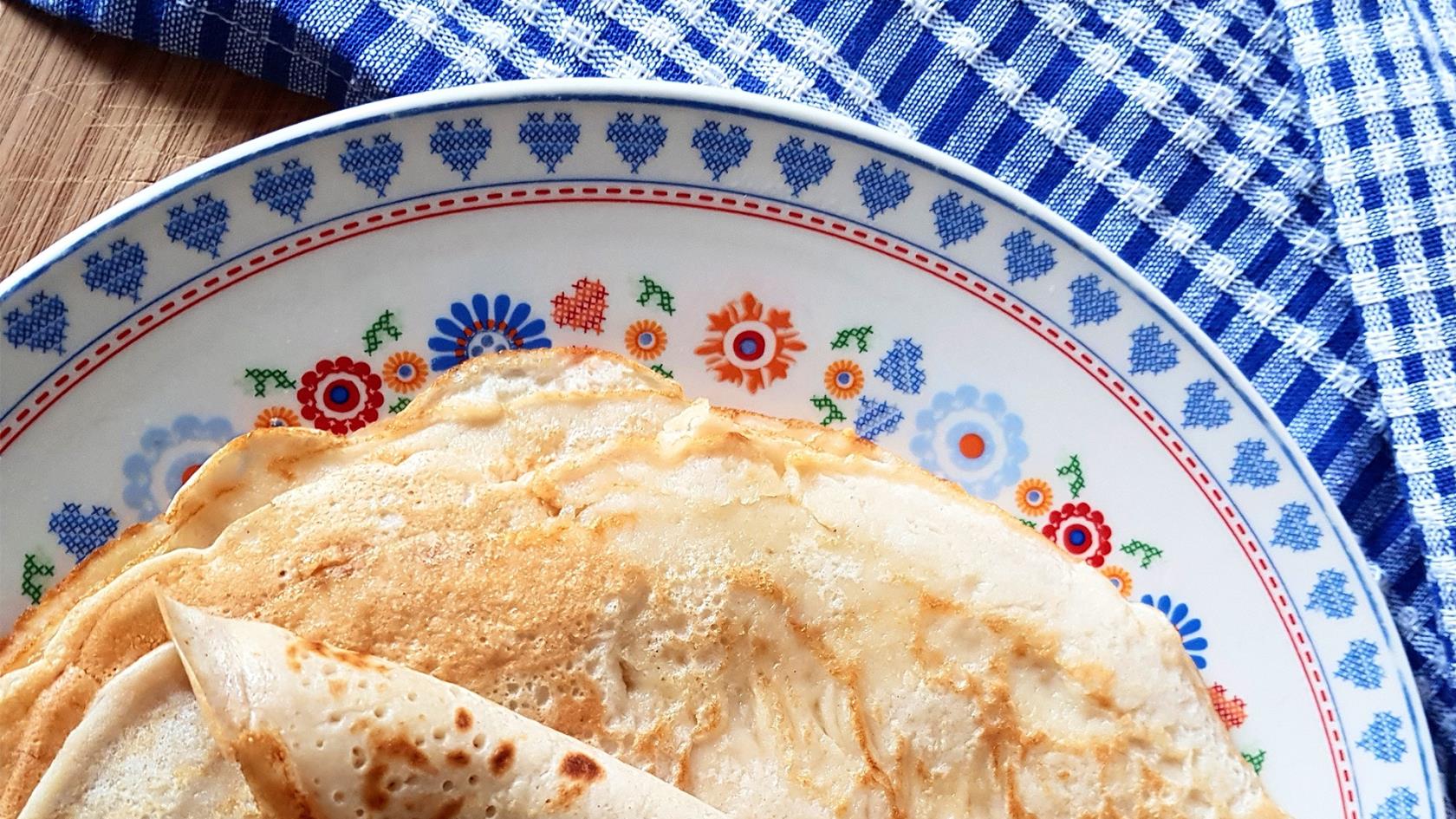 Pannkakor i hög på en blommig porslinstallrik på bord med blåvitrutig duk