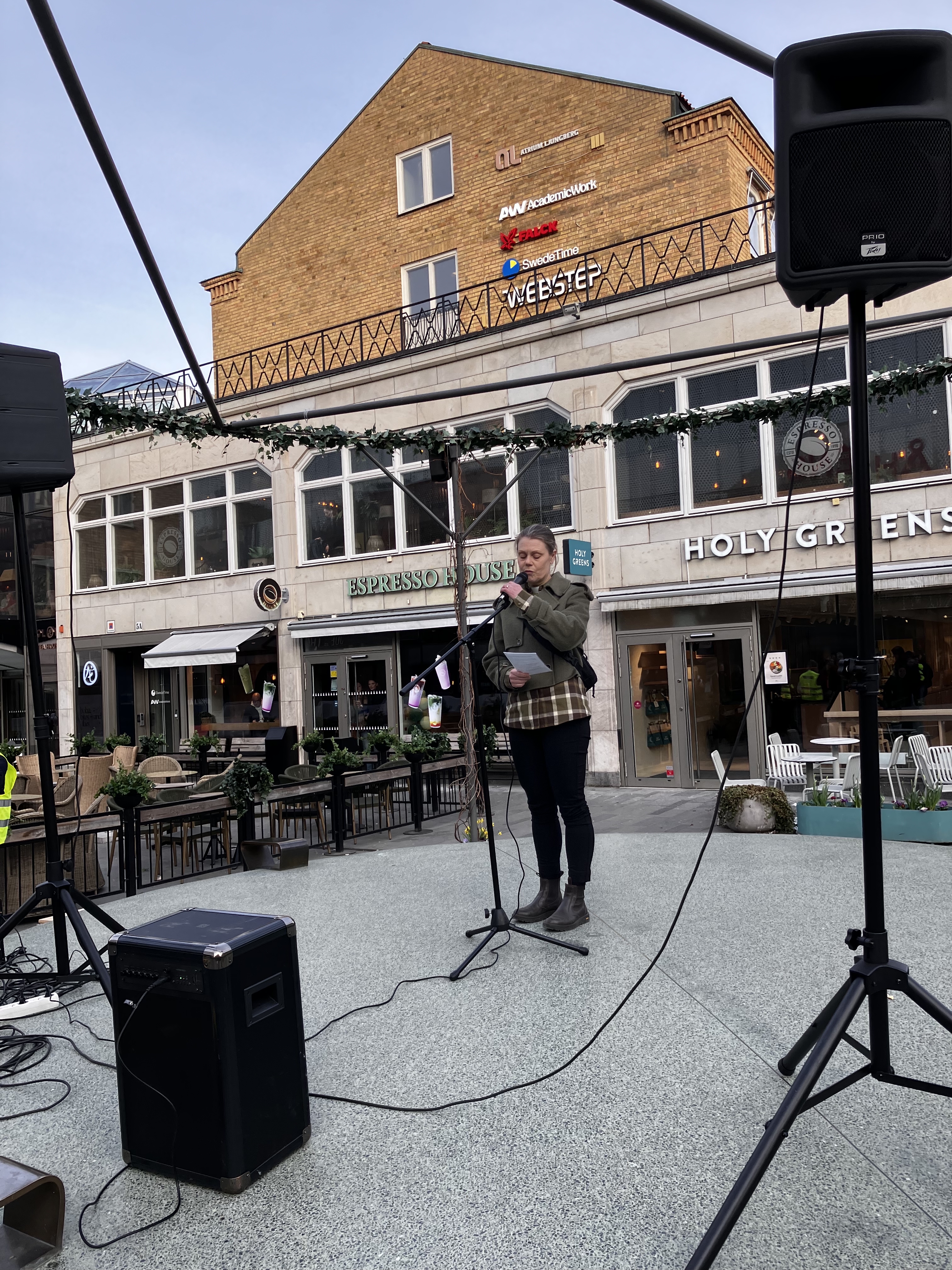 En kvinna står framför en mikrofon på ett torg och talar.