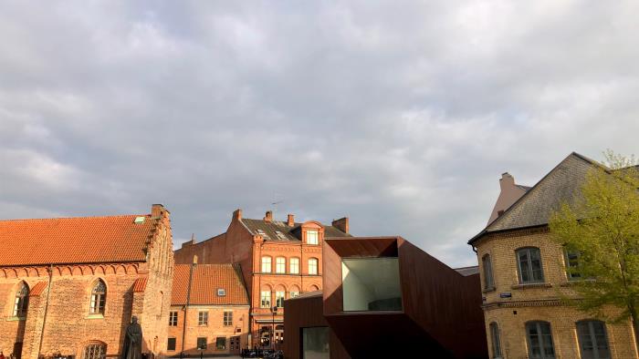 Ett torg med två röda tegelbyggnader, en modern byggnad i corten och en gul tegelbyggnad. På torget står en staty och det finns en fontän.