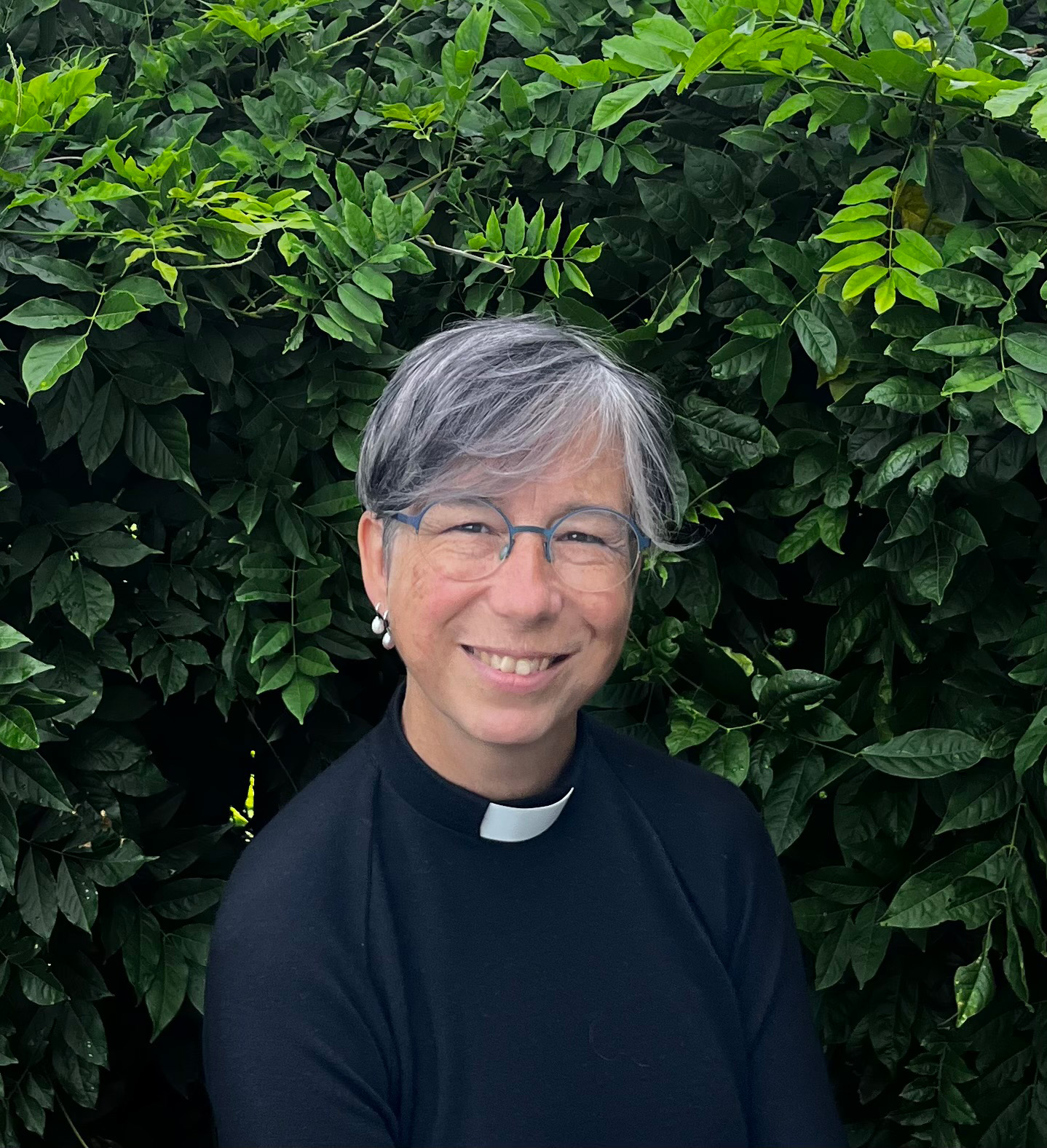 Leende svartklädd kvinna i frimärksskjorta framför ett buskage.