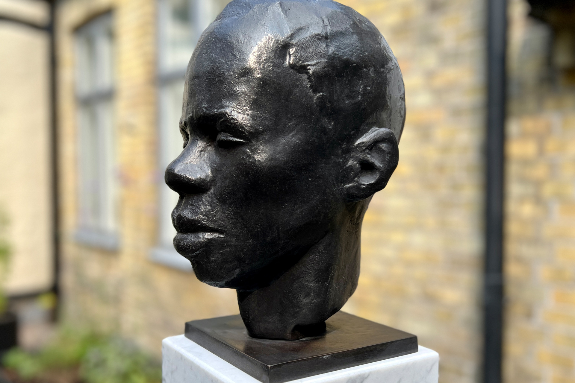 Byst av brons på ett marmorfundament framför en gul tegelvägg.