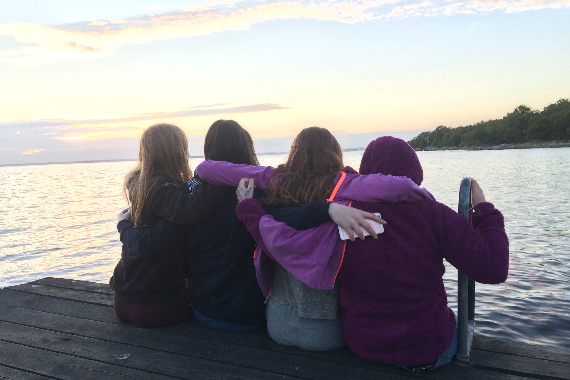 Fyra ungdomar sitter på en brygga och håller varandra om ryggarna. Framför bryggan syns en sjö. 