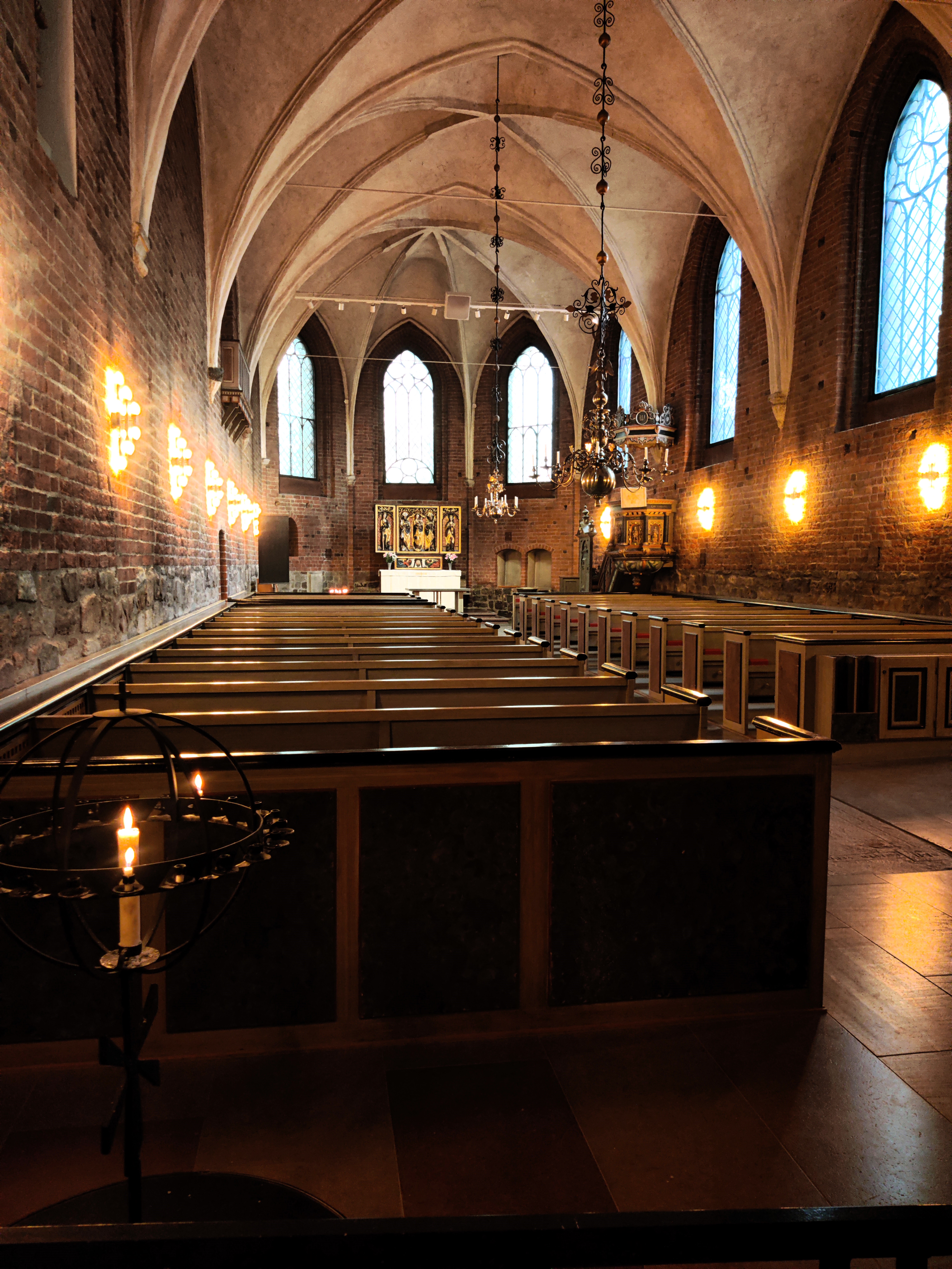 Bild tagen inne i kyrkan. Längst bort syns koret med sin altartavla som är belyst. I förgrunden syns ljusbäraren samt bänkraderna.   