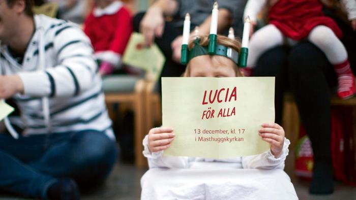 En liten lucia sitter på golvet och läser i ett papper. På pappret står det Lucia för alla.