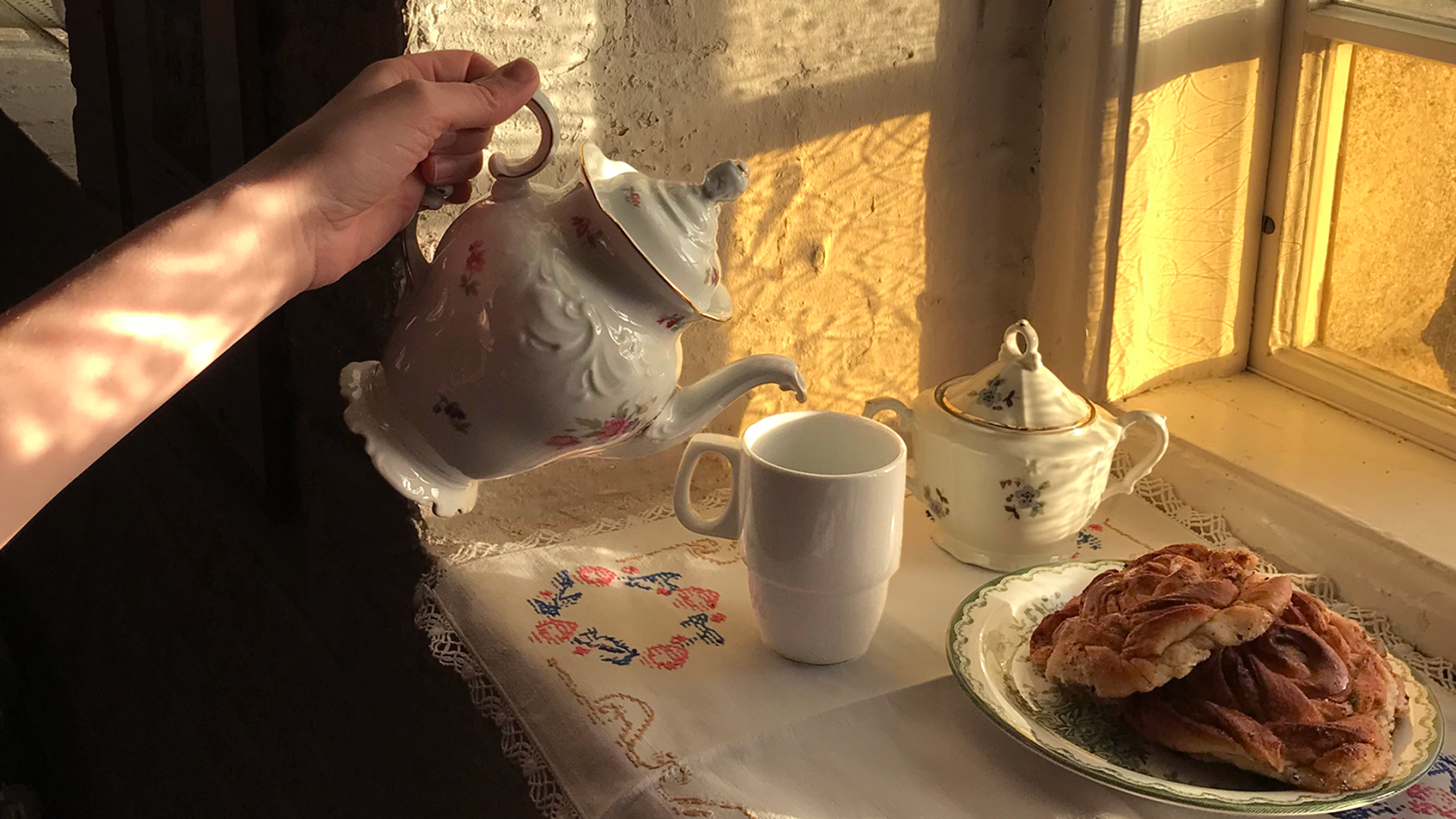 Någon häller upp te från en gammal engelsk tekanna i en tekopp. Bredvid står ett fat med kanelbullar.