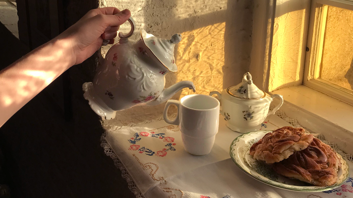 Någon häller upp te från en gammal engelsk tekanna i en tekopp. Bredvid står ett fat med kanelbullar.