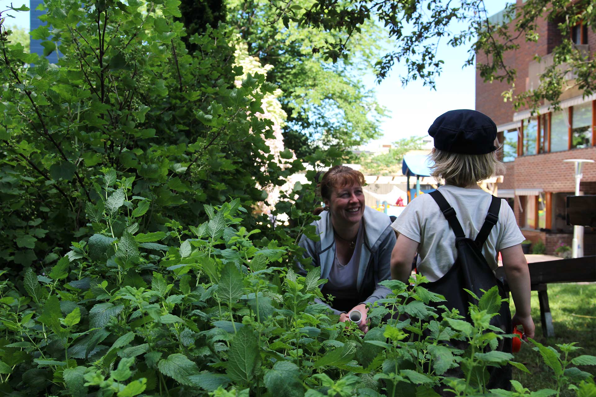 Ett barn på förskolan leker i buskarna på gården. En av pedagogerna sitter på huk och ler mot barnet.