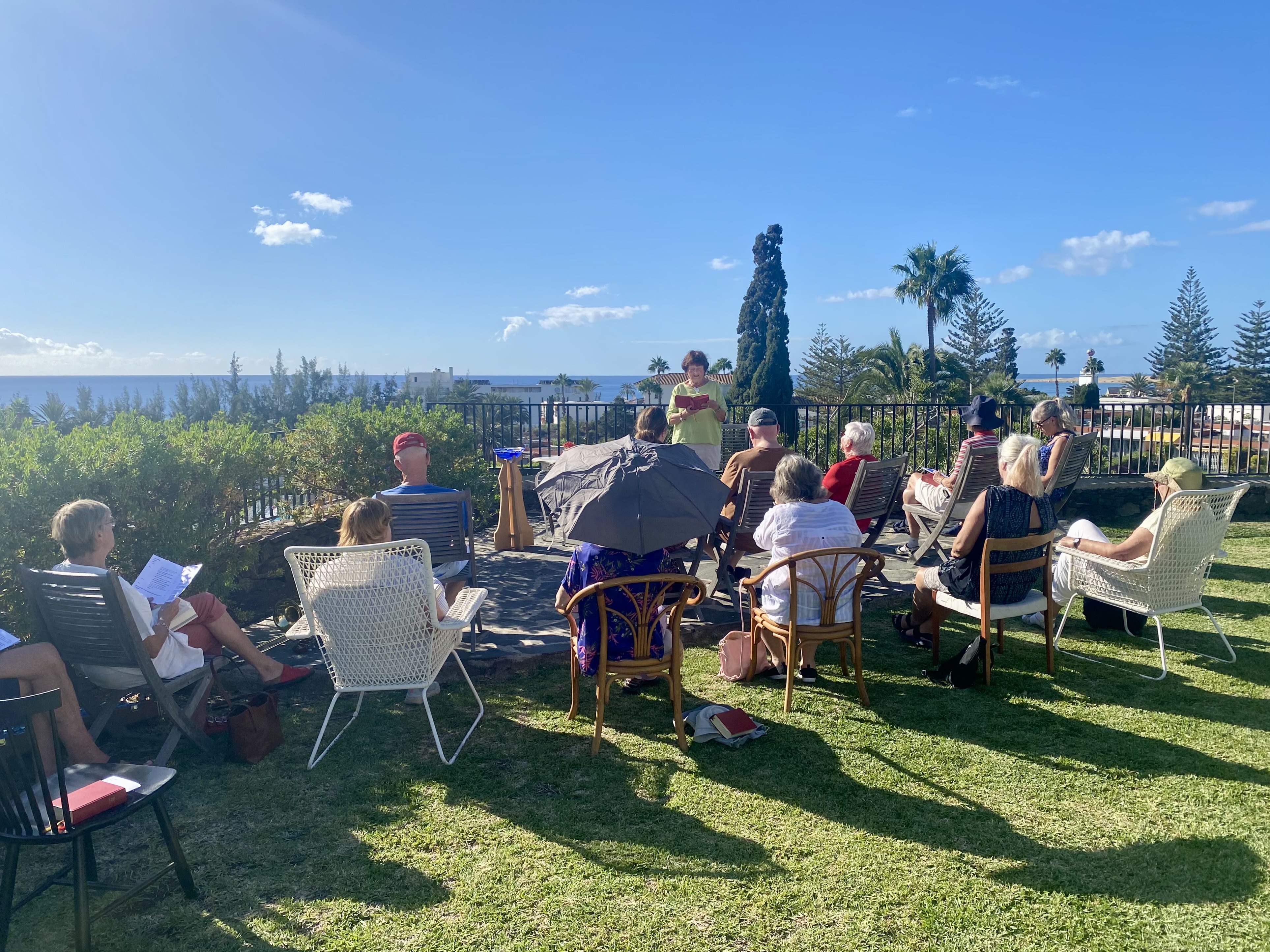 En grupp människor är samlade till andakt i Prästgårdsparken. Solen lyser och både himlen och havet är blått.