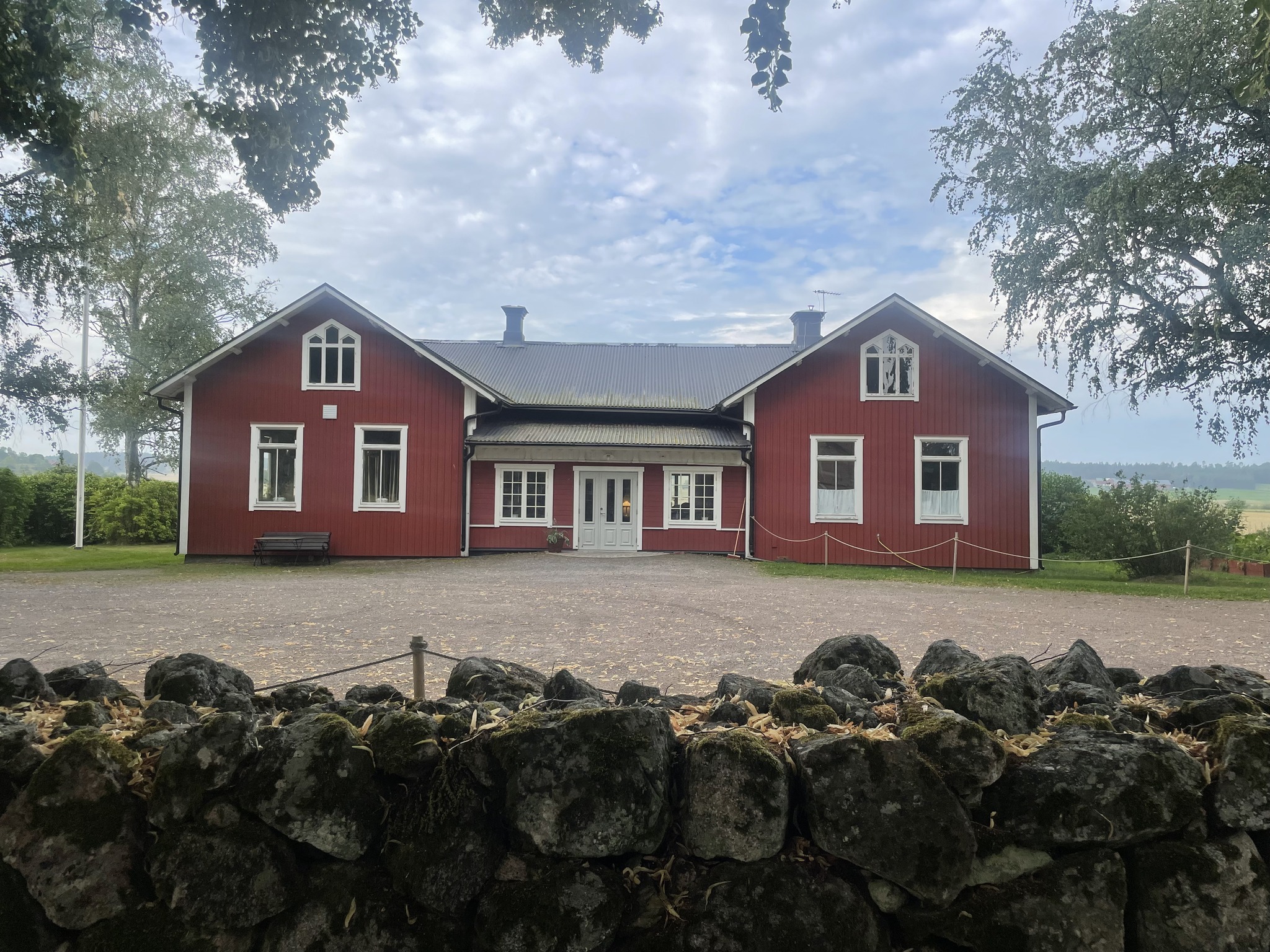 Nysätras gamla kyrkskola, ett rött trähus, är centrerat i bilden. I förgrunden syns muren till Nysätra kyrka.