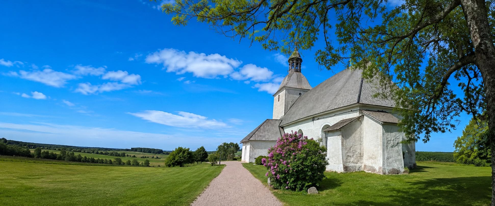 Vit kyrka med högt torn och grått tak, omgiven av grönskande träd. Himlen är klarblå i solsken.