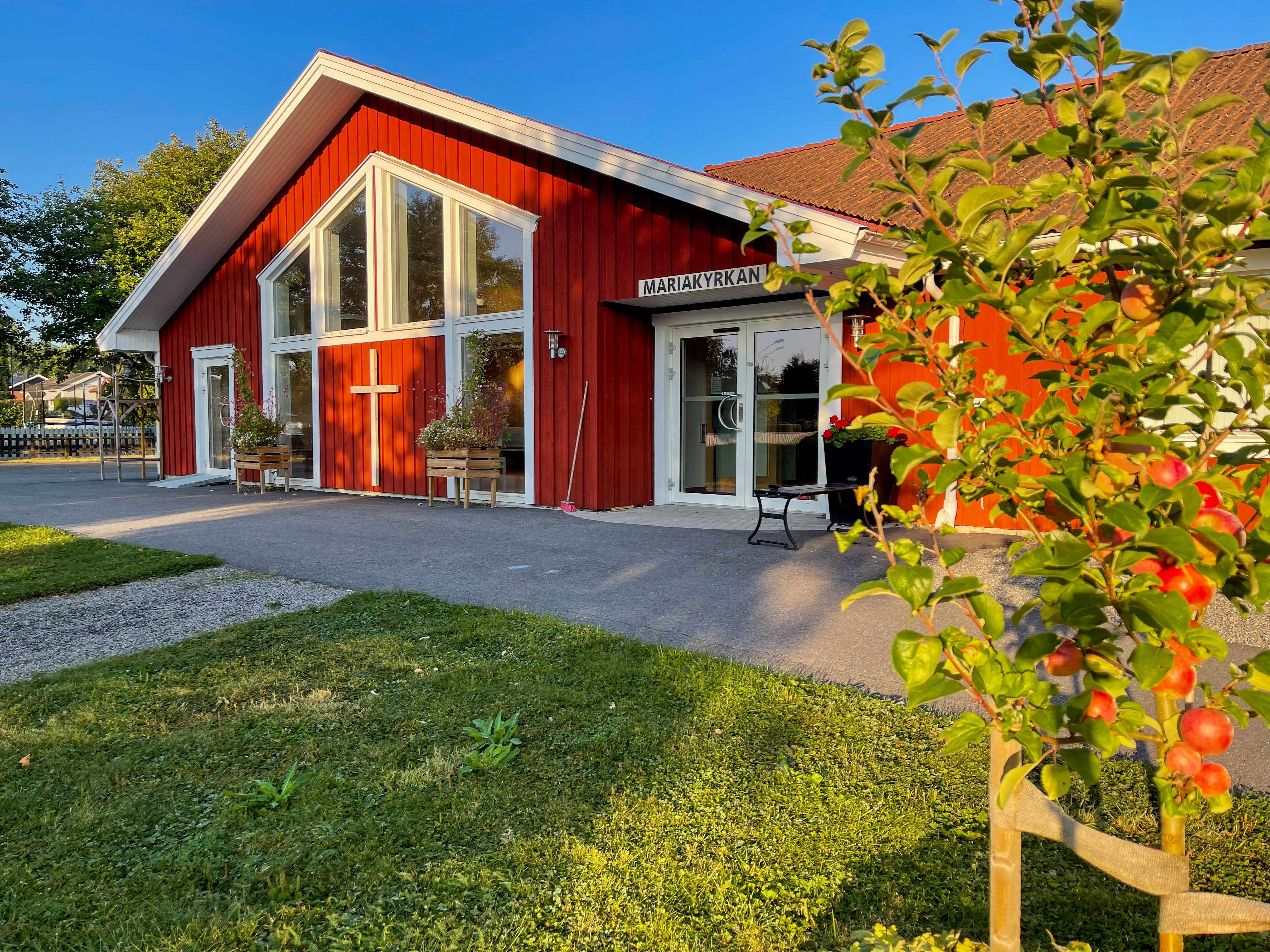 Röd träkyrka med vita knutar och stora fönster. Vid entrén syns ett kors och skylten ’Mariakyrkan’. I förgrunden står ett litet äppelträd med röda äpplen i solsken.