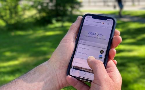 Närbild på händer som håller en mobiltelefon och är inne på en sida på nätet där man kan boka dop.