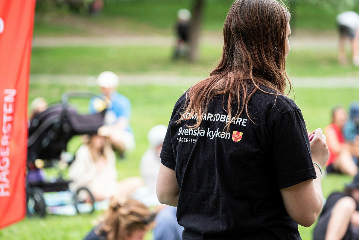 En ung kvinna står med ryggen mot kameran i en park där föräldrar och barn sitter framför henne i gräset. På hennes svarta t-shirt står det Sommarjobbare.