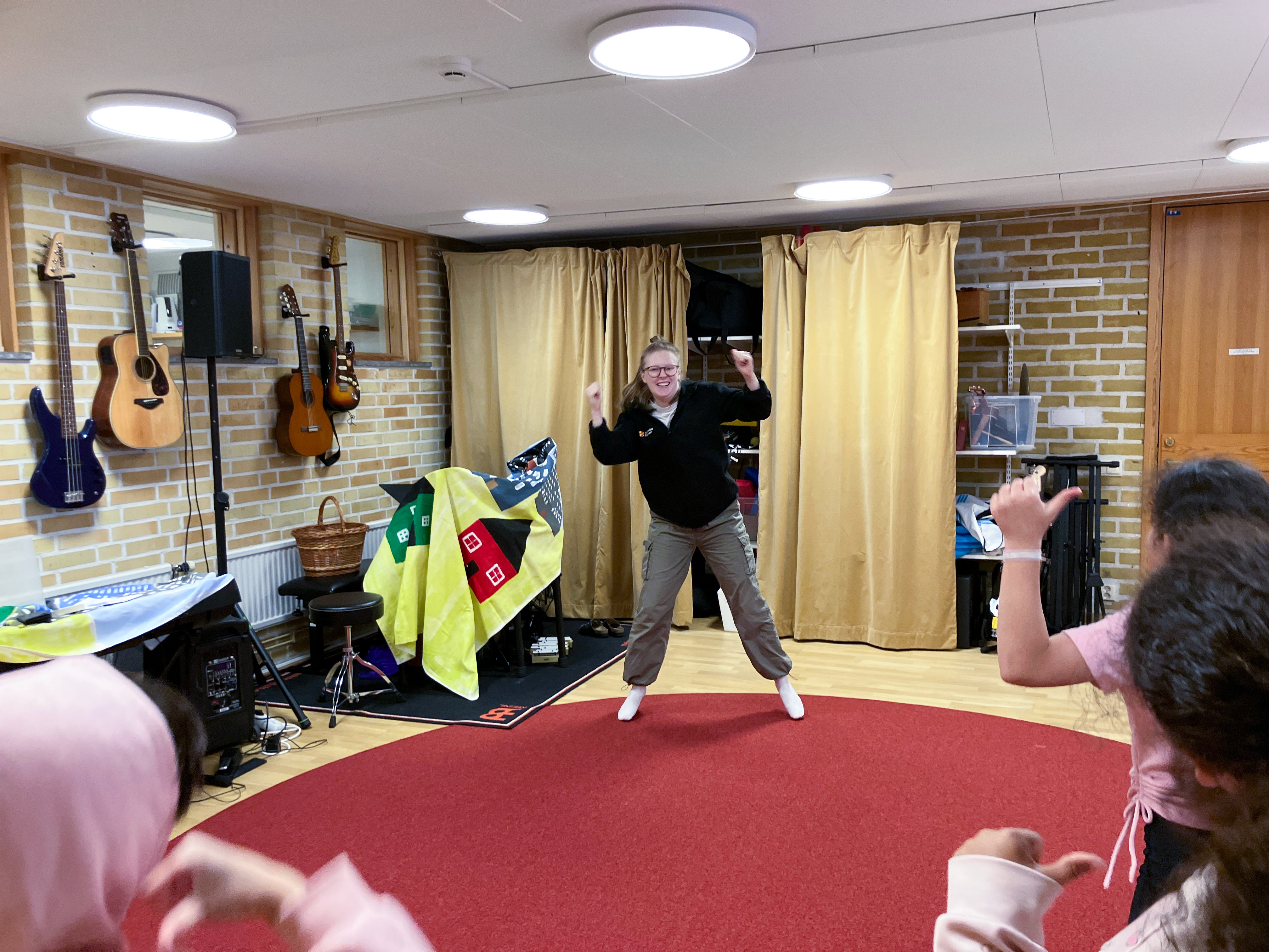 En dansledare dansar i ett musikrum och tre barn, sedda bakifrån, härmar rörelsen.