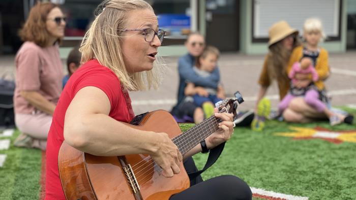 Kvinna spelar gitarr. I bakgrunden sitter några mammor och barn.