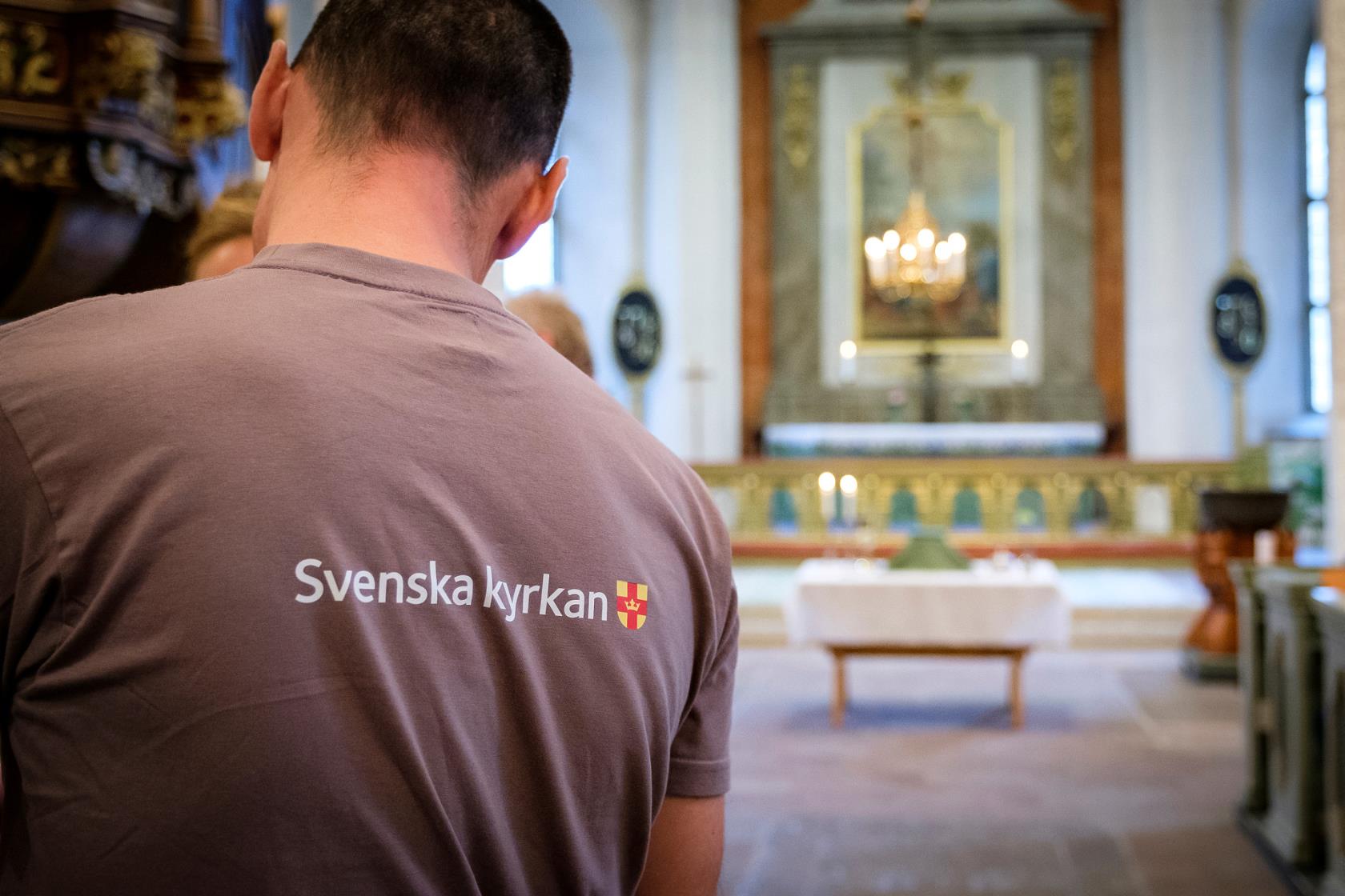 Person i grå T-shirt med Svenska kyrkan-logga i ett kyrkorum