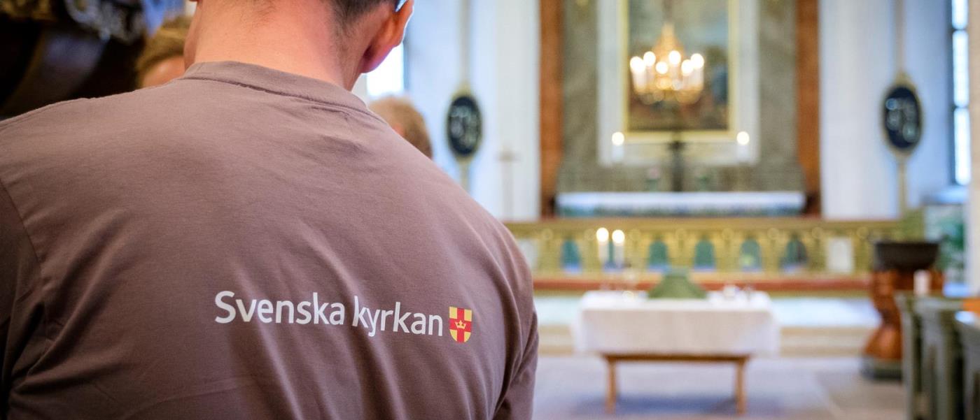 Person i grå T-shirt med Svenska kyrkan-logga i ett kyrkorum