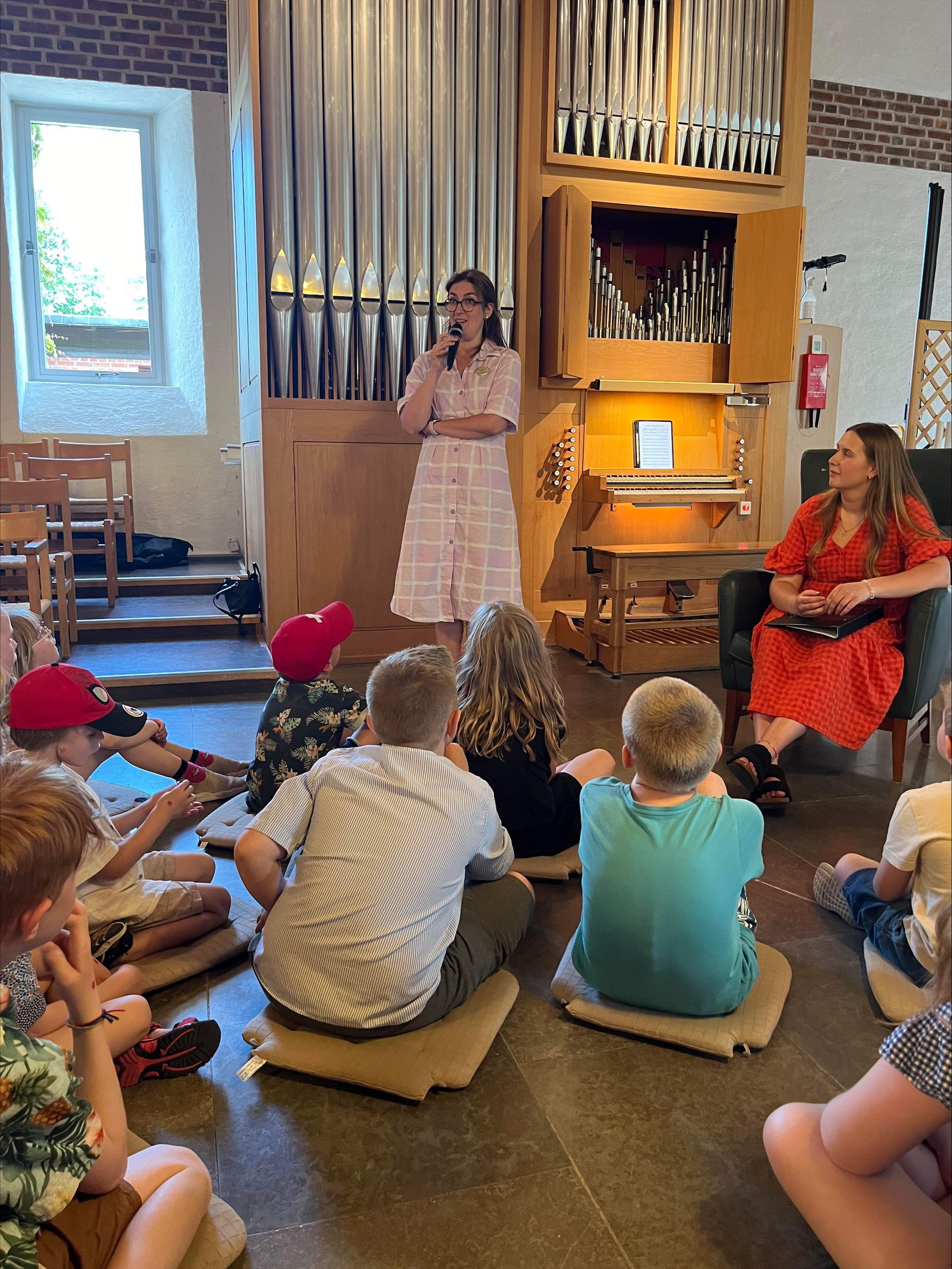 En grupp barn framför en orgel som lyssnar på Maria och Cecilia som berättar ett orgeläventyr