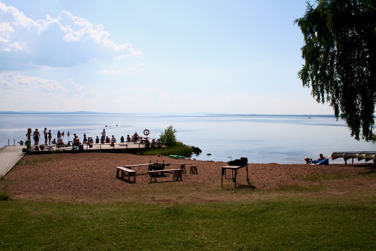 Vy över sandstranden vid Stiftsgården Rättvik med brygga där massor med folk befinner sig.