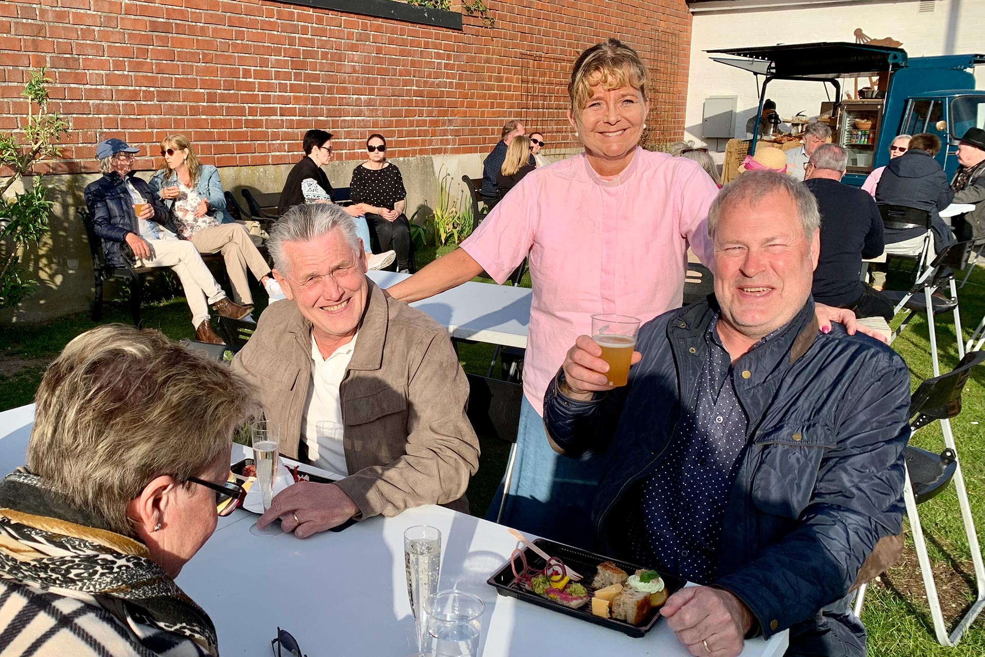 Två män sitter och äter och dricker vid ett bord. Bakom står en kvinna.