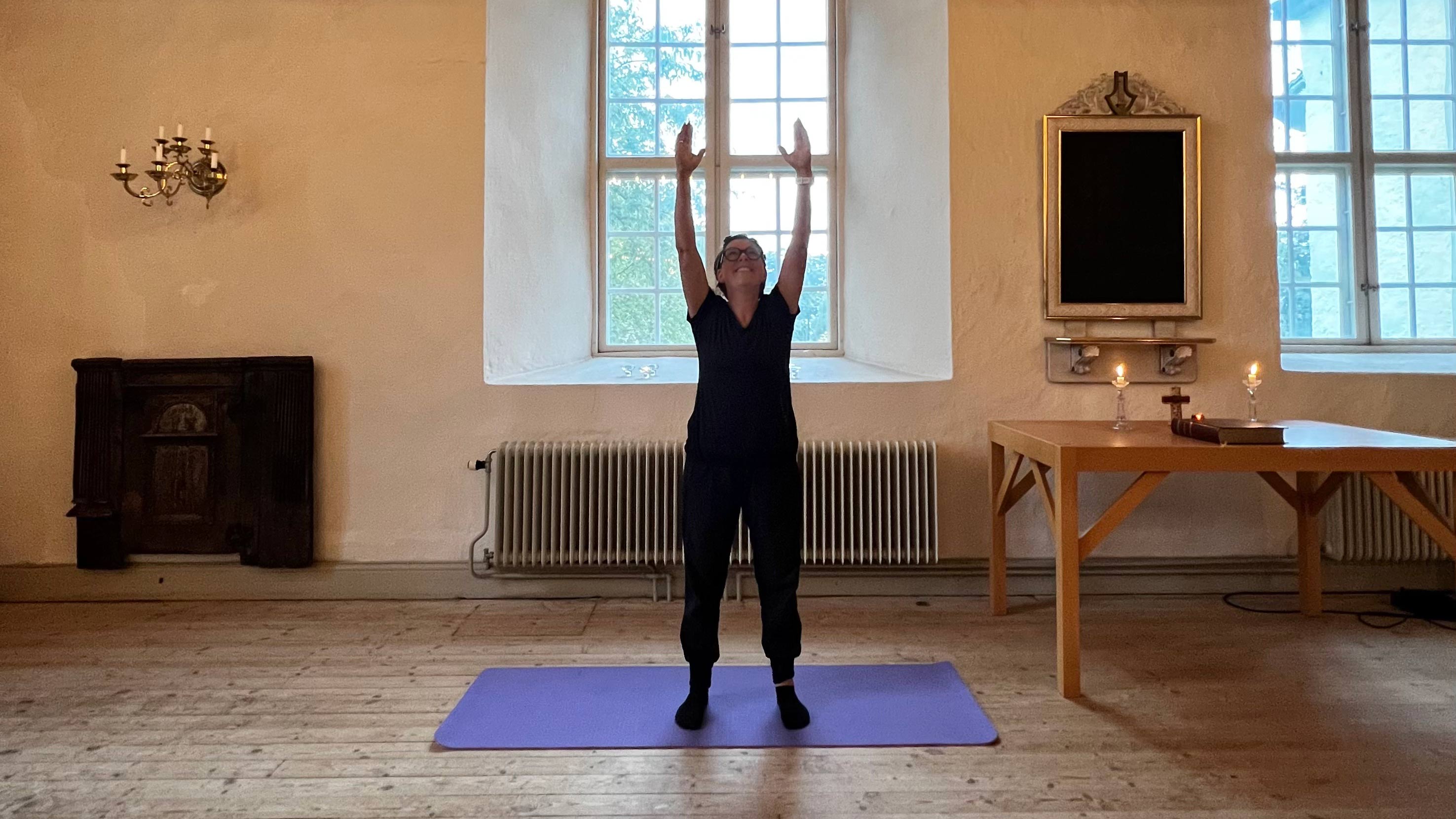 Kvinna står på yogamatta i ett kyrkorum och har händerna sträckta mot taket.