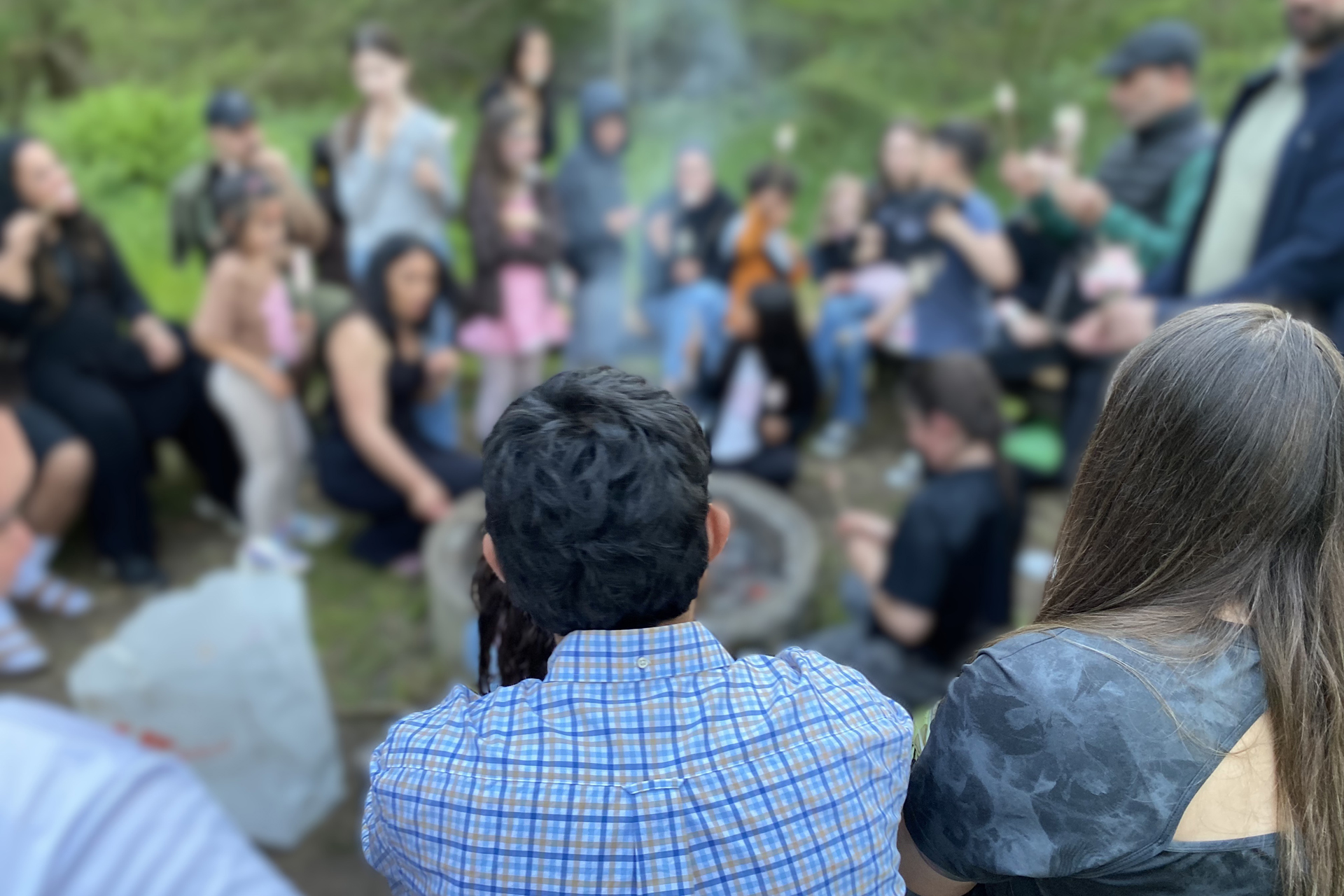 Tre personer sitter vid en grill med ryggen mot kameran. Framför dem anas fler människor men de är suddiga.