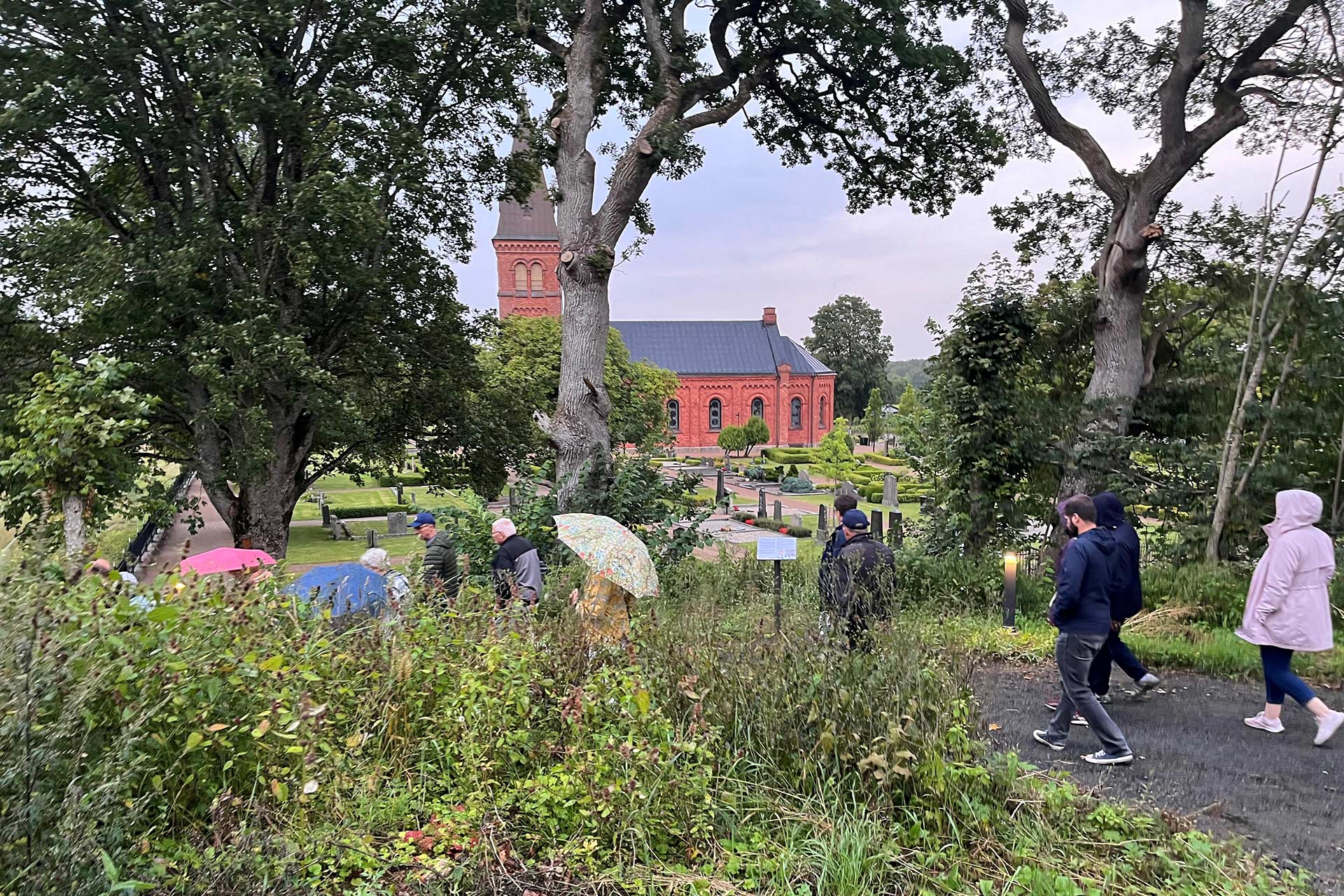 Människor med regnkappor och paraply promenerar längs en grusstig. I bakgrunden syns en röd tegelkyrka.