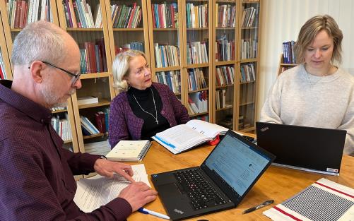 En man och två kvinnor sitter runt ett bord och arbetar med två laptops, papper och pärm.