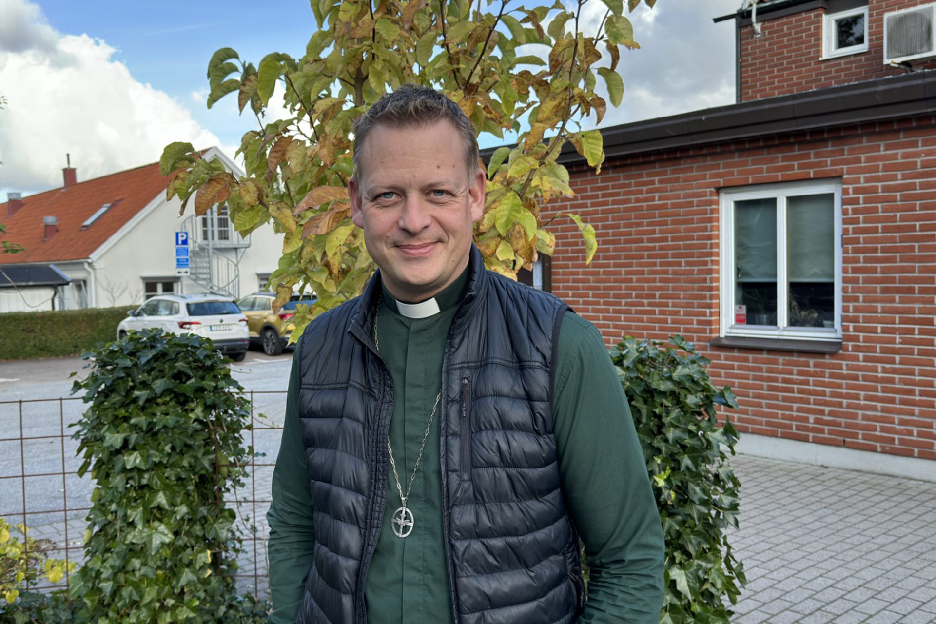 Man i diakonskjorta och svart täckväst står utanför ett hus.