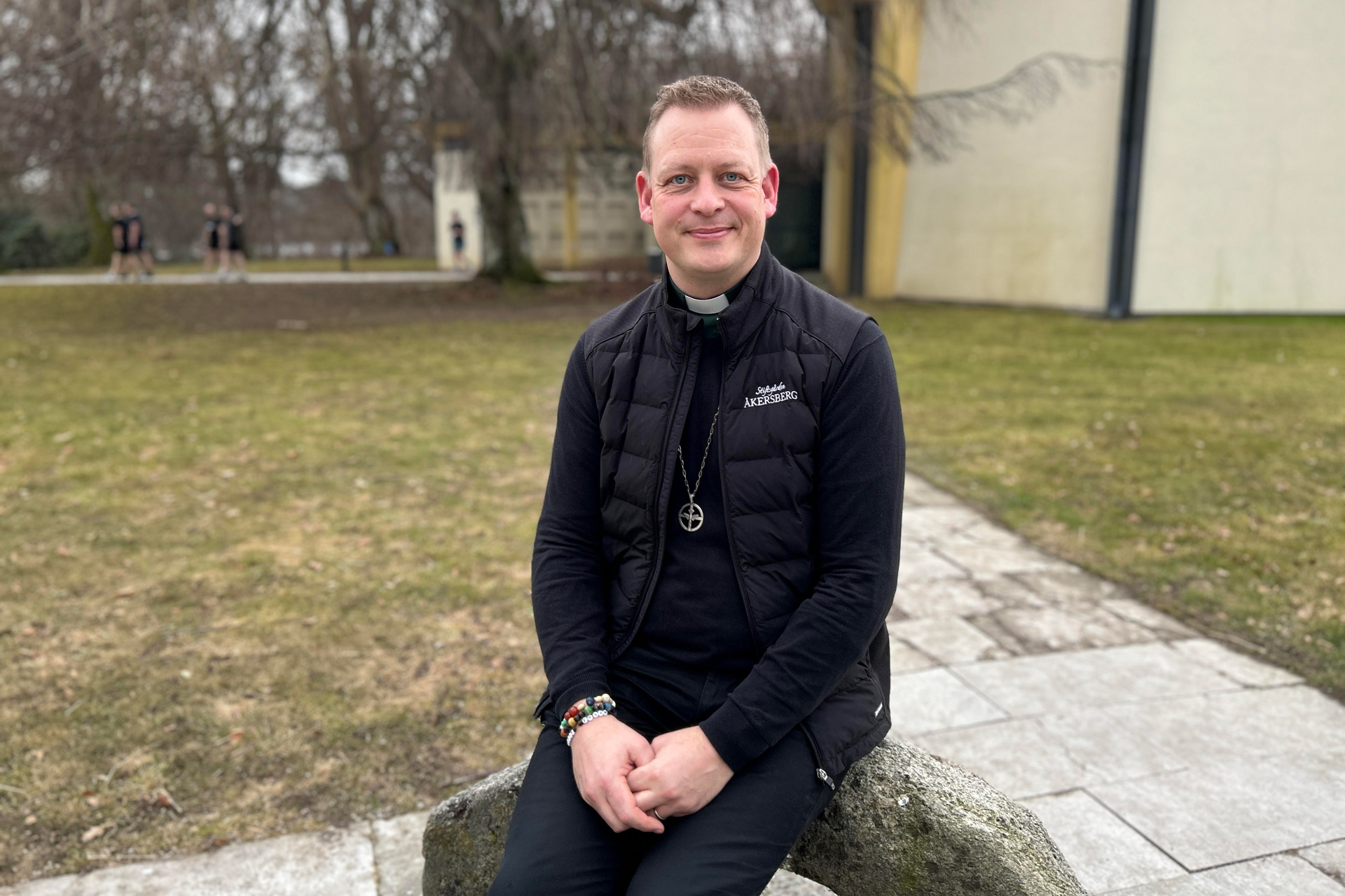 En man sitter på en sten. Bakom honom syns en byggnad, ett stort träd och gröna gräsmattor. Runt halsen bär han ett halsband. Det runda hänget föreställer en duva i ett kors.