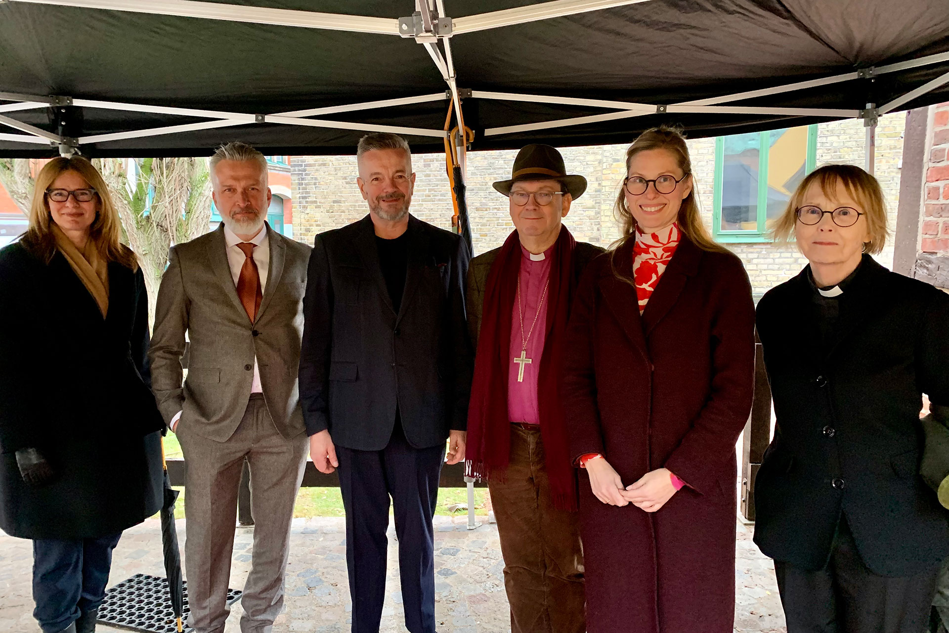 Tre män och tre kvinnor står utomhus under ett tälttak.