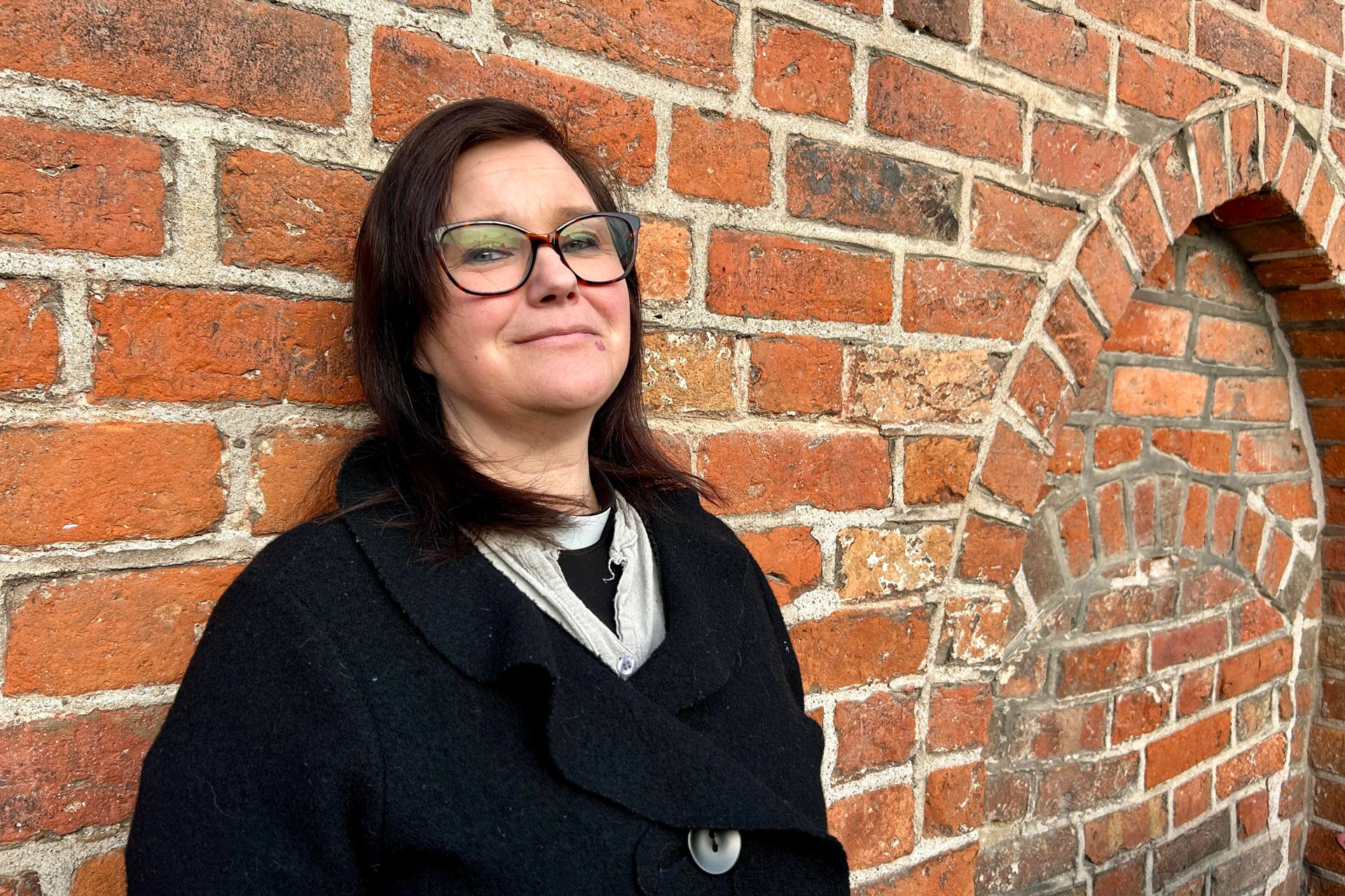 En kvinna med axellångt mörkt hår och glasögon står lutad mot en röd tegelvägg. På väggen finns ett murat valv. Hon är klädd i svarta kläder och prästskjorta.