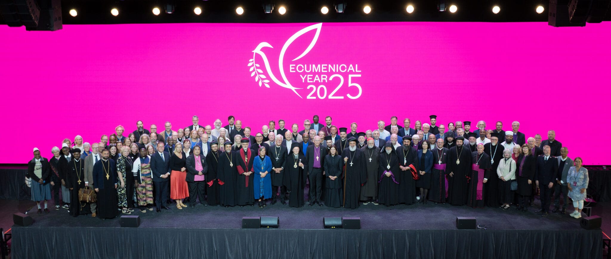 En stor folksamling poserar glatt framför en lila skärm med loggan för ekumeniska året 2025.