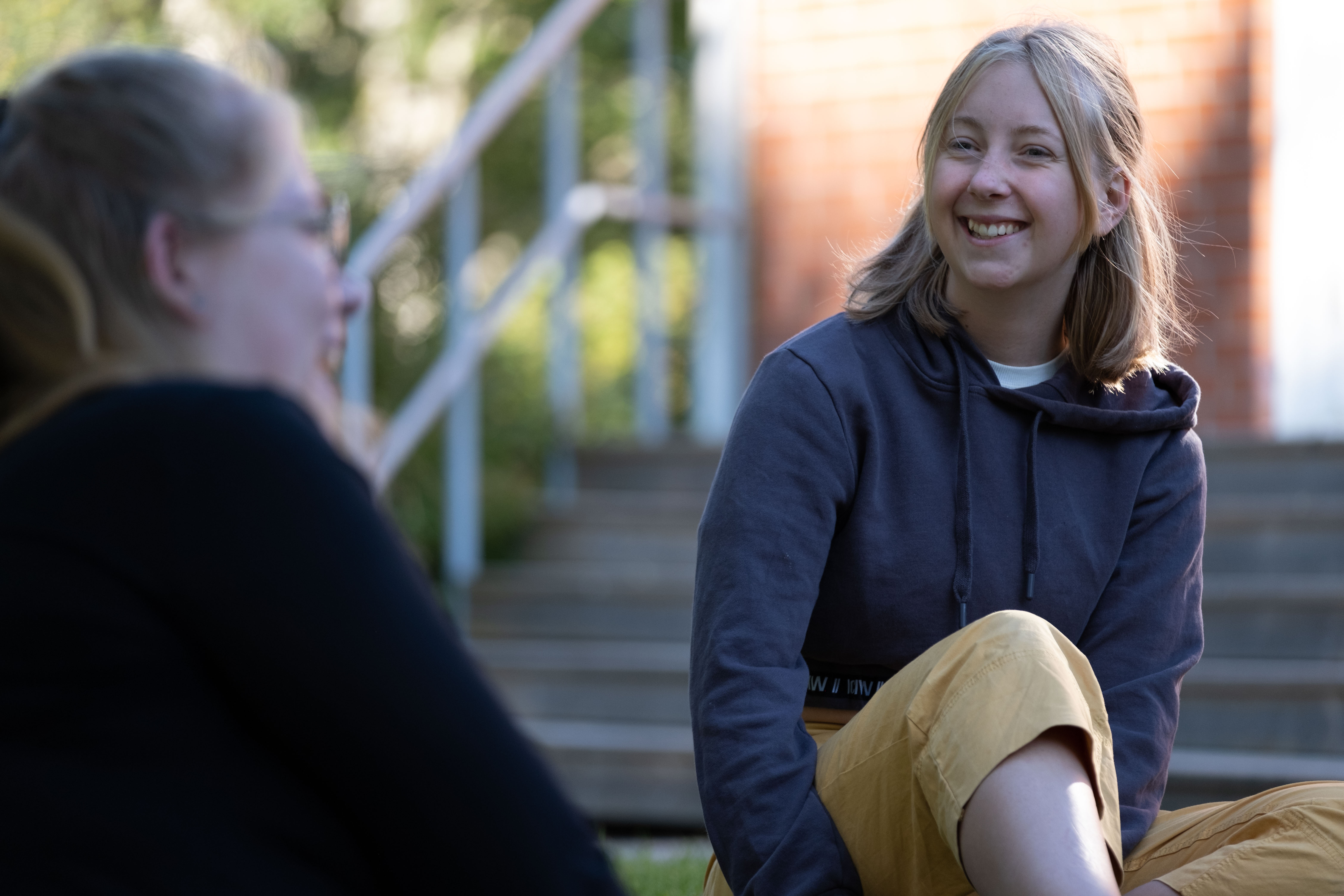 En ung kvinna sitter i utomhus en solig dag och ler åt en vän i förgrunden.