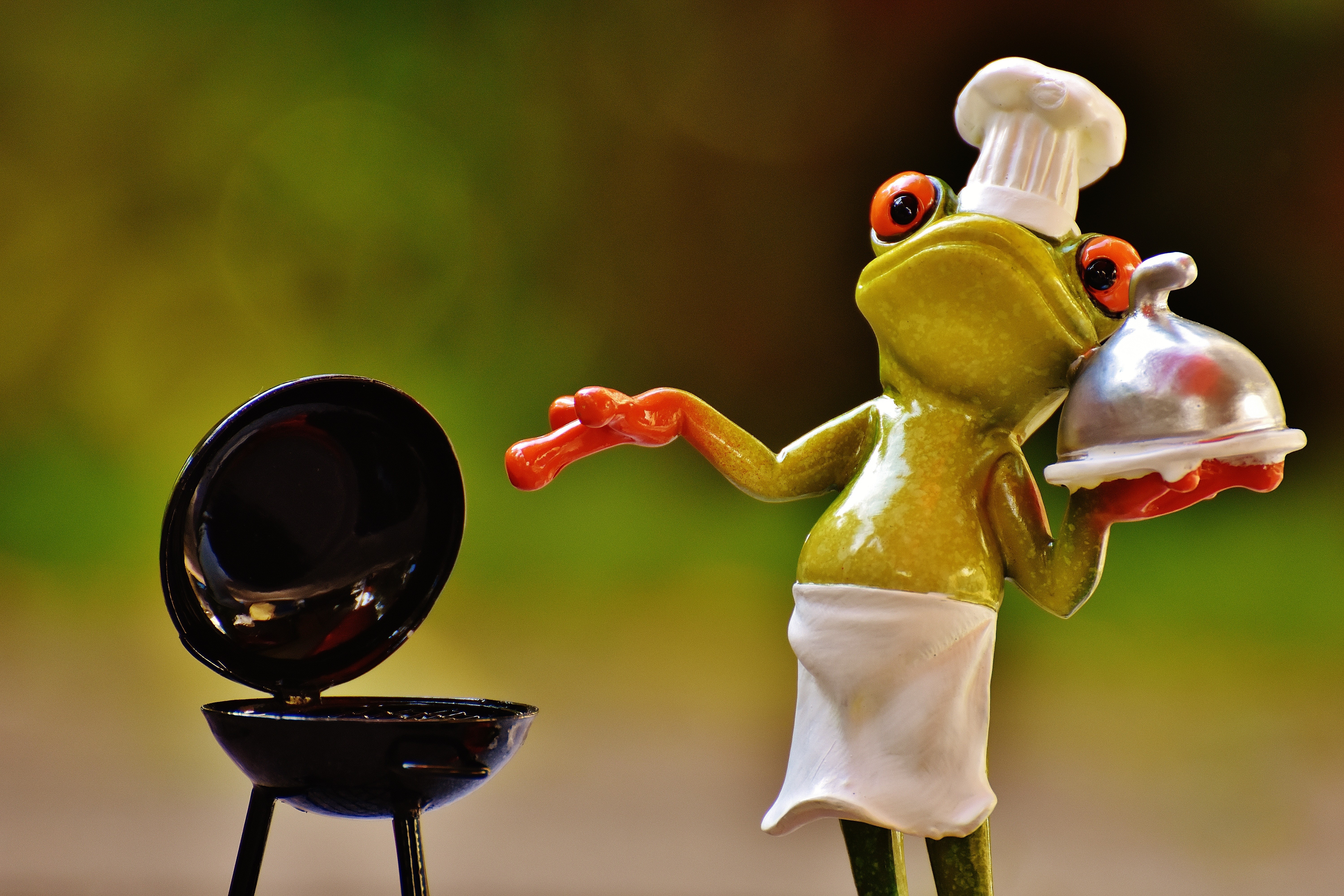 Bild med en groda i kockmössa och förkläde står vid en liten grill med öppet lock