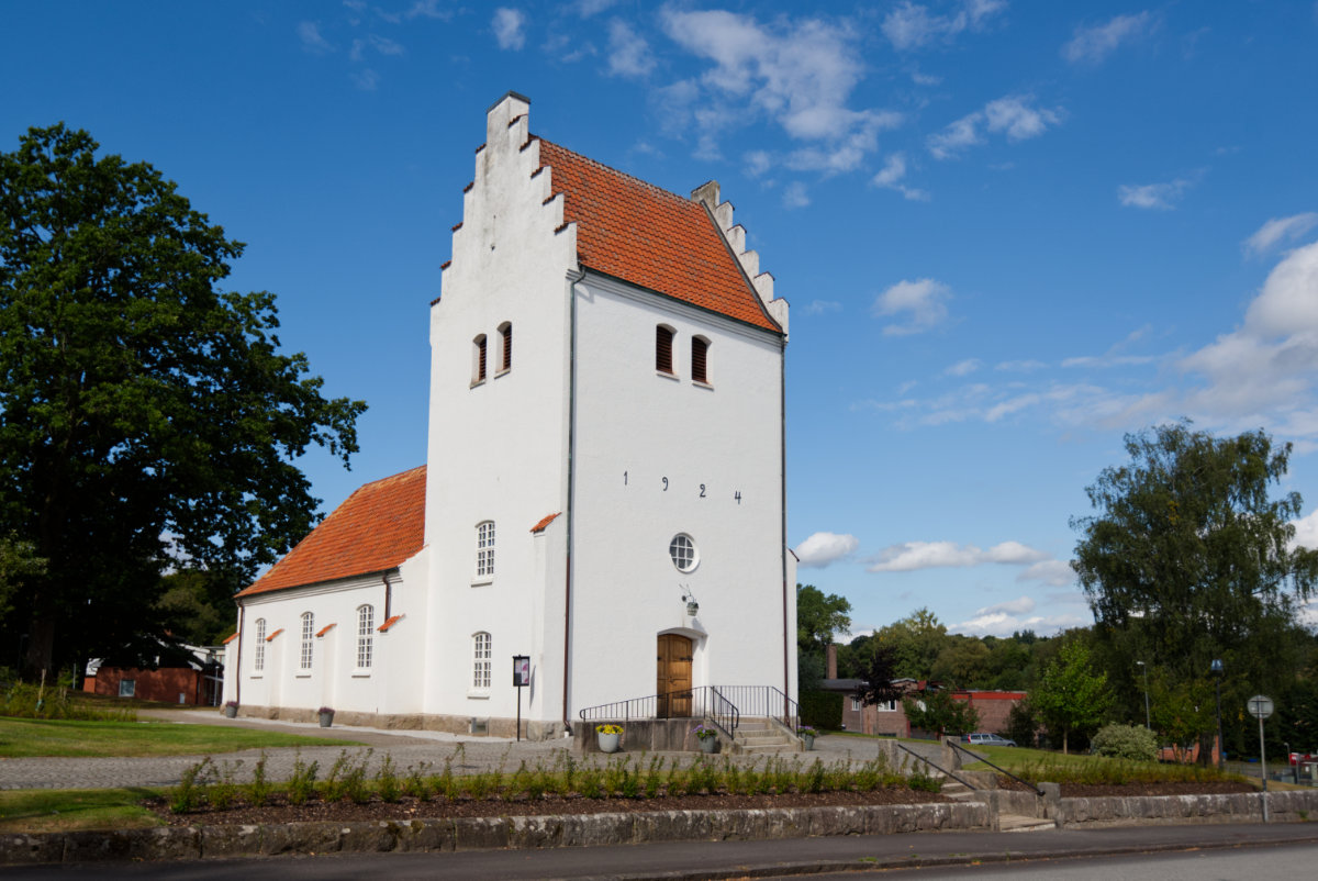 En mindre, vit kyrka med klocktorn som vätter ut mot en gata. Kyrkans tegeltak är orangefärgat. Himlen är blå med några vita moln.