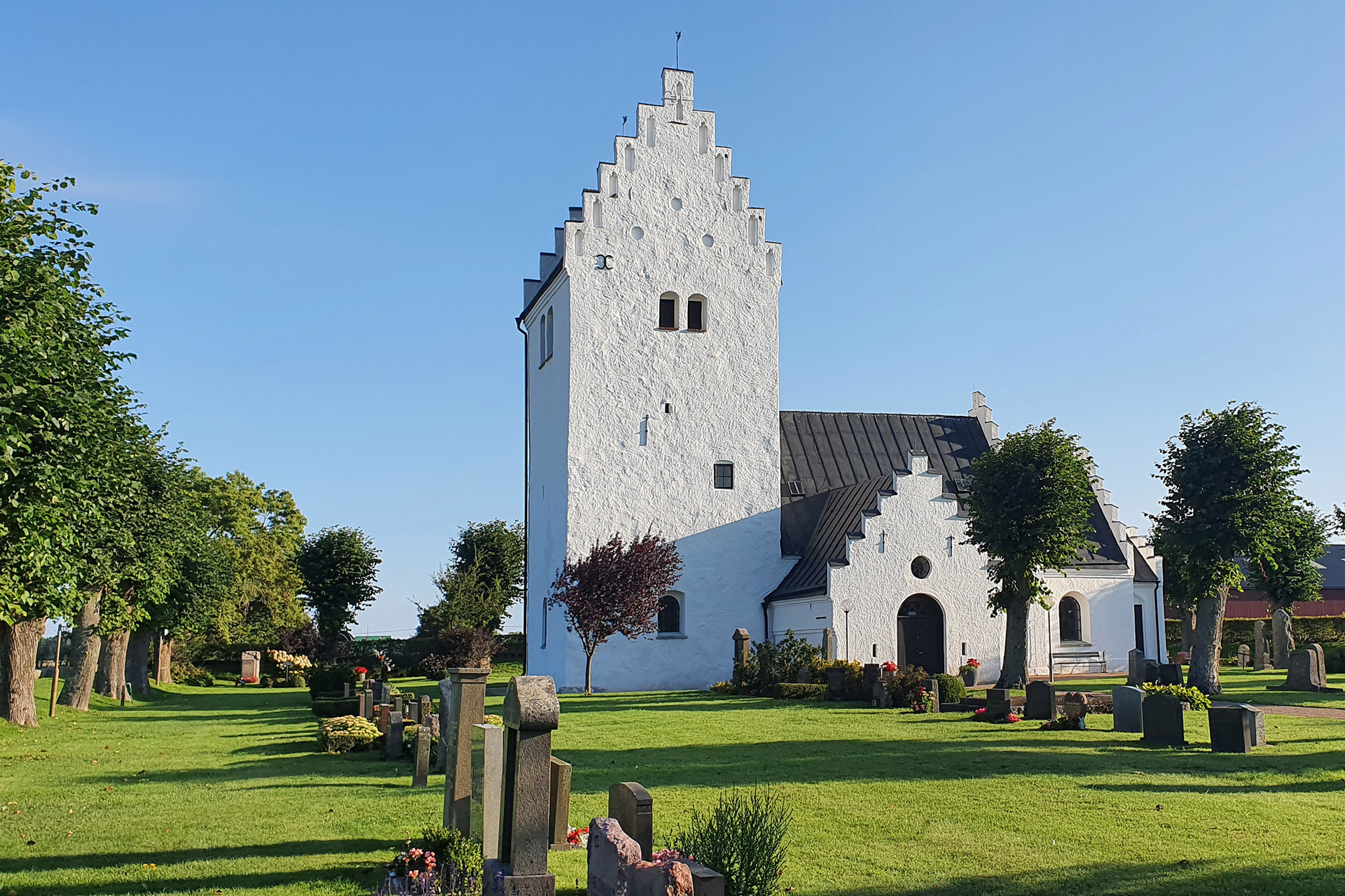 Vit kyrka med grönskande kyrkogård framför belyst av sensommarsol