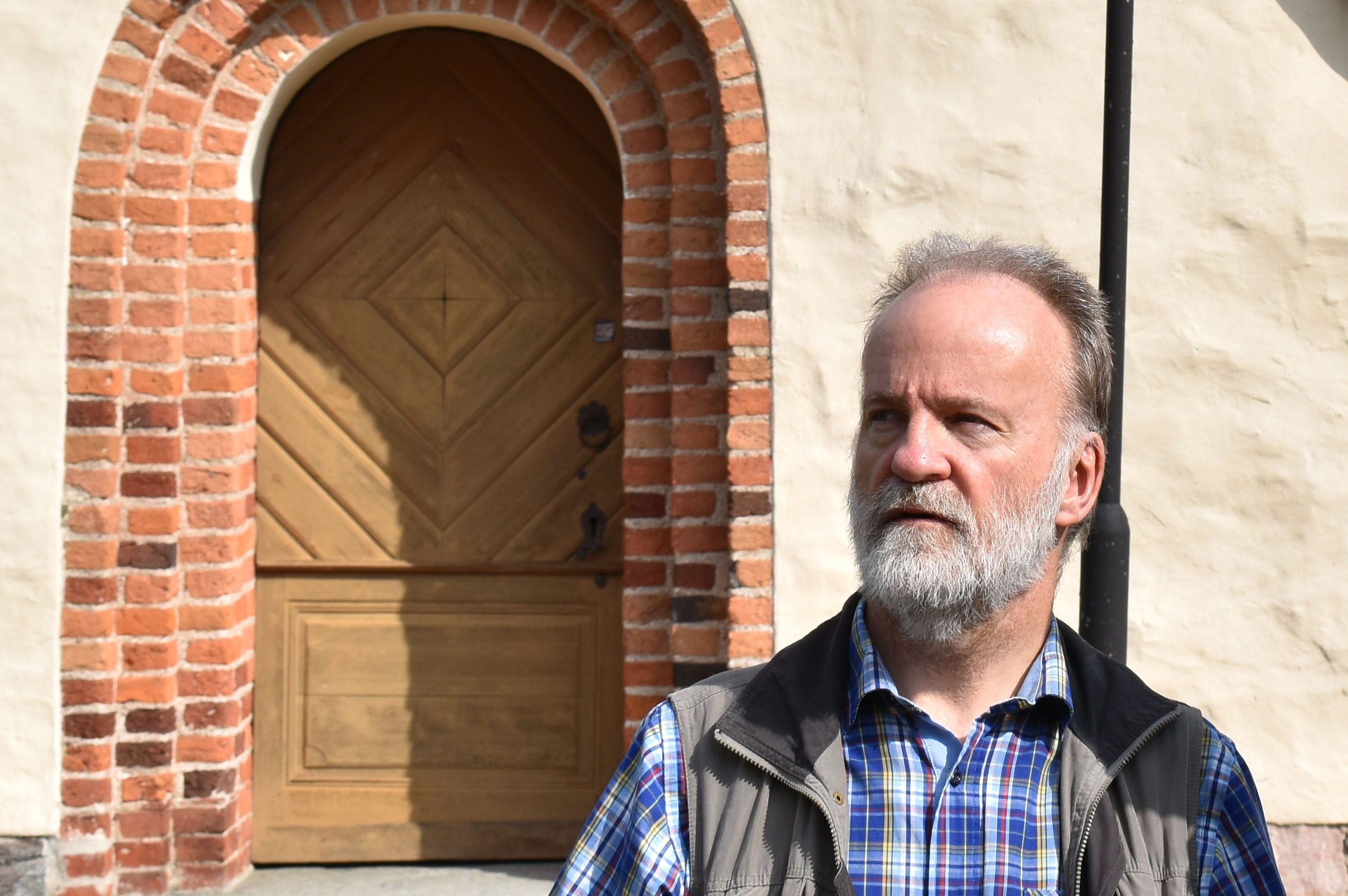 Man med grått skägg framför kyrka.